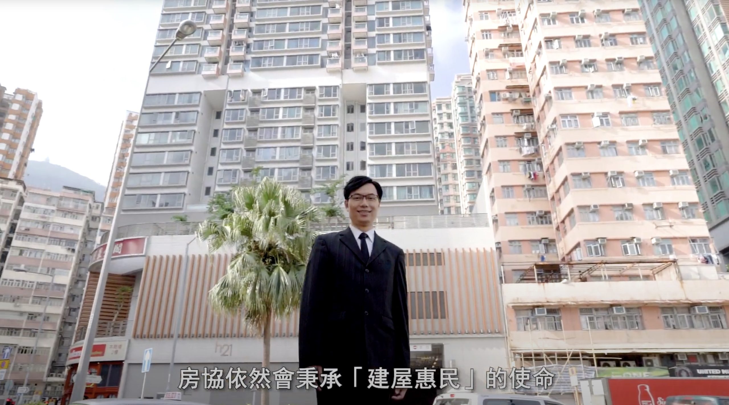 A man in a suit stands smiling in front of tall apartment buildings in an urban setting. There is a palm tree beside him. Chinese subtitles appear at the bottom of the image.