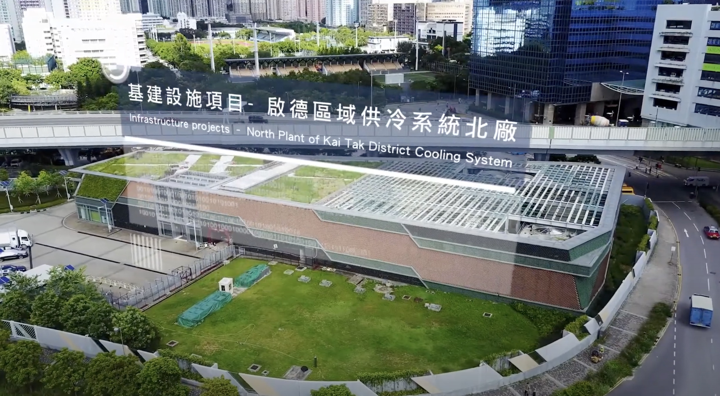A modern building with a green rooftop labeled “North Plant of Kai Tak District Cooling System,” surrounded by grass, trees, and nearby city buildings. Large bilingual signage in Chinese and English is visible on the roof.