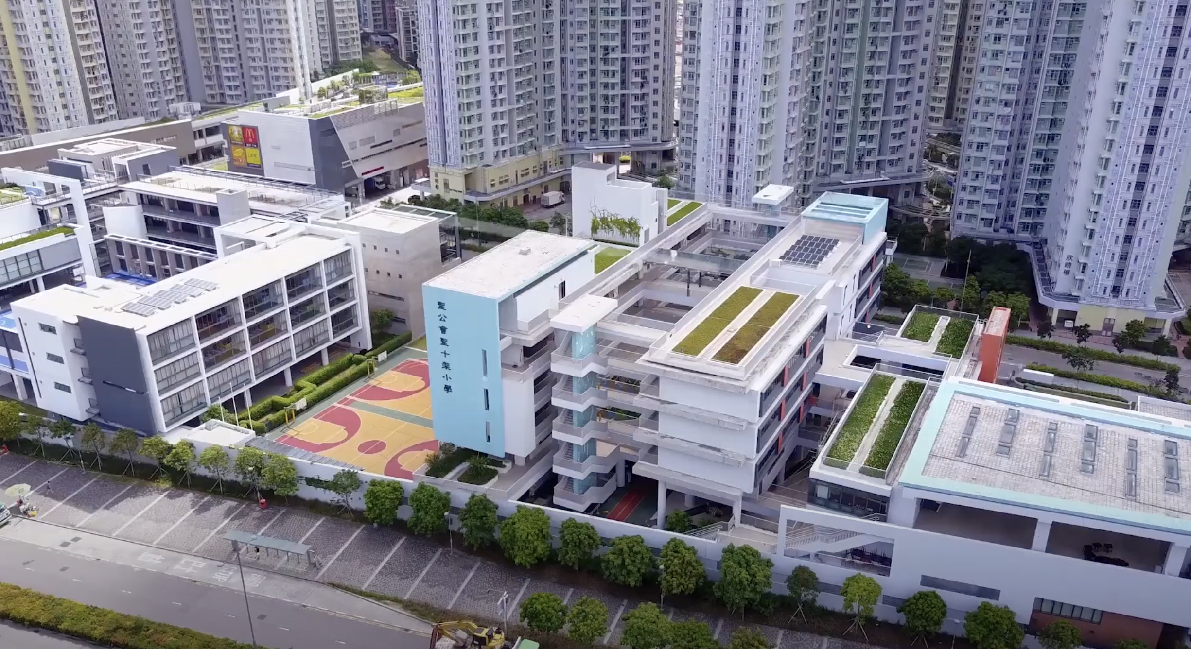 A modern school complex with several white multi-story buildings, green roofs, and a colorful outdoor basketball court, surrounded by high-rise apartment buildings in an urban area.