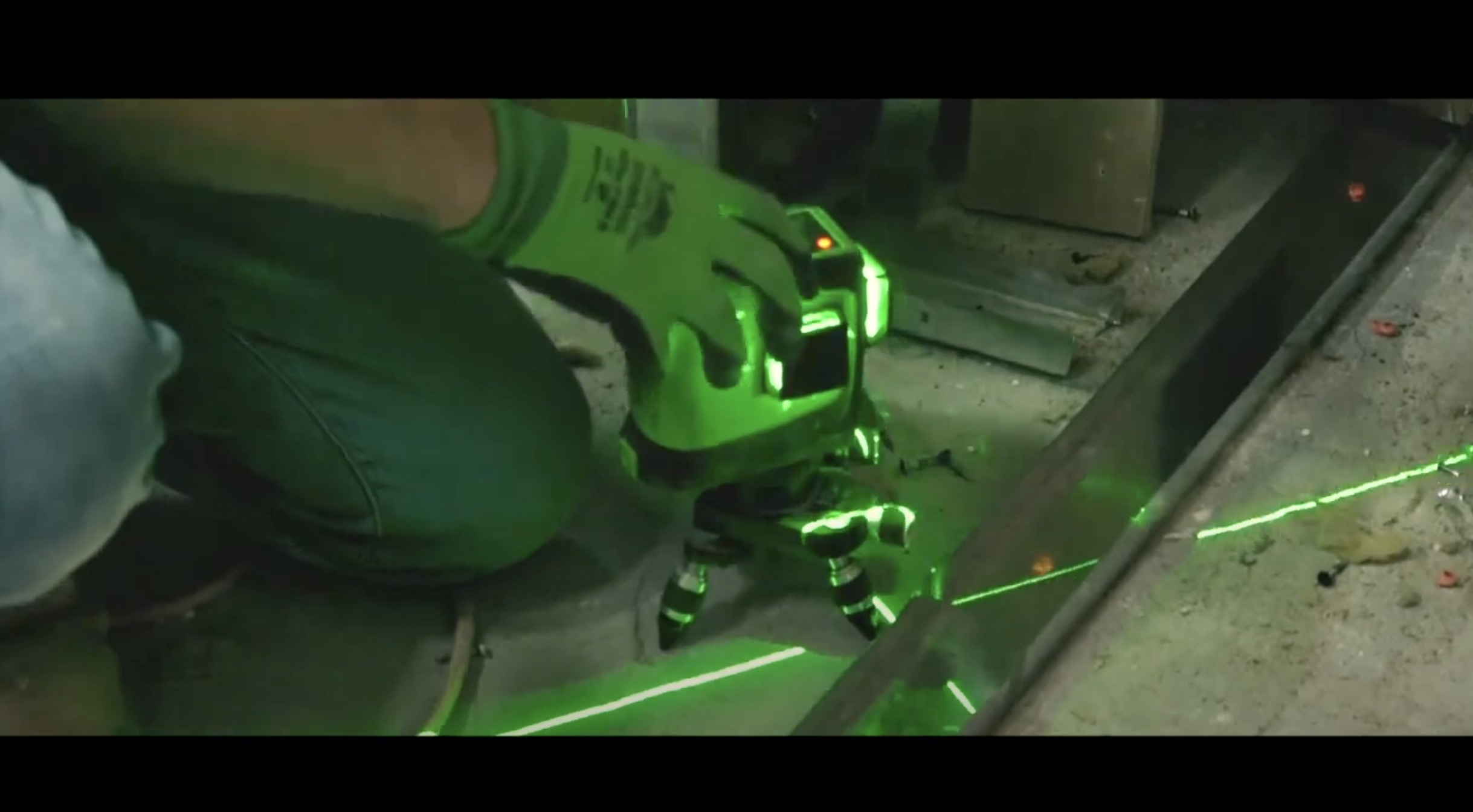 A person wearing green gloves adjusts a laser level tool on a concrete surface, with bright green laser lines projecting across the floor and metal rails.