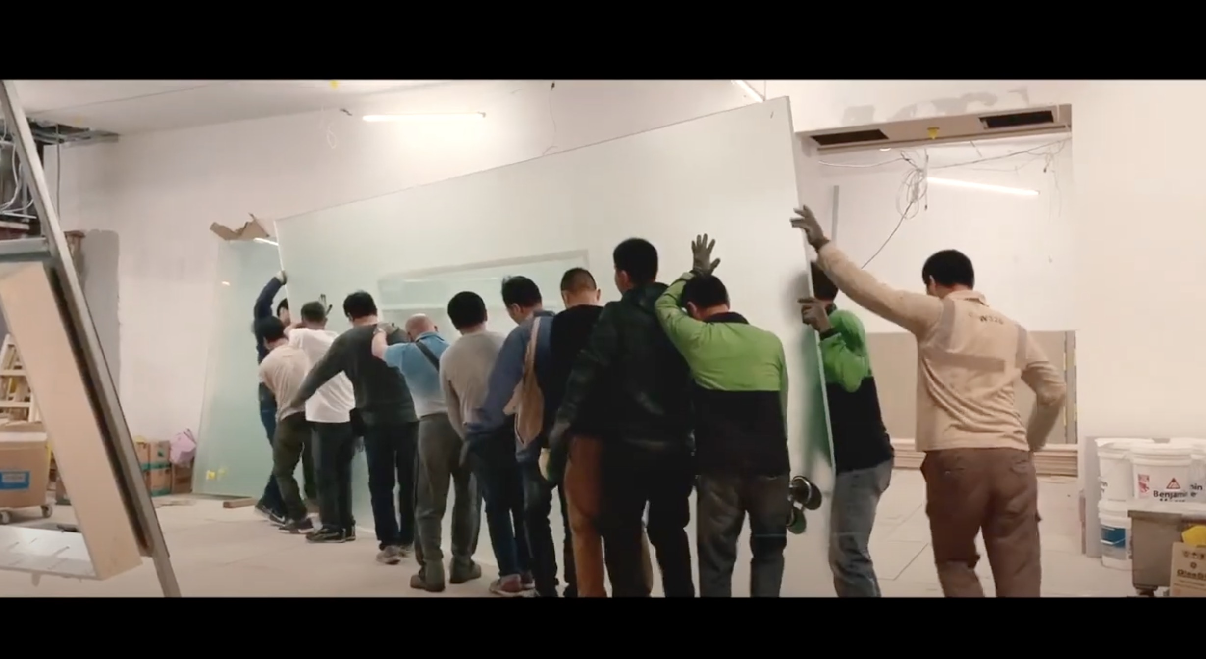 A group of construction workers carefully lifts and moves a large glass panel inside a building under construction, working together to maneuver it into place.