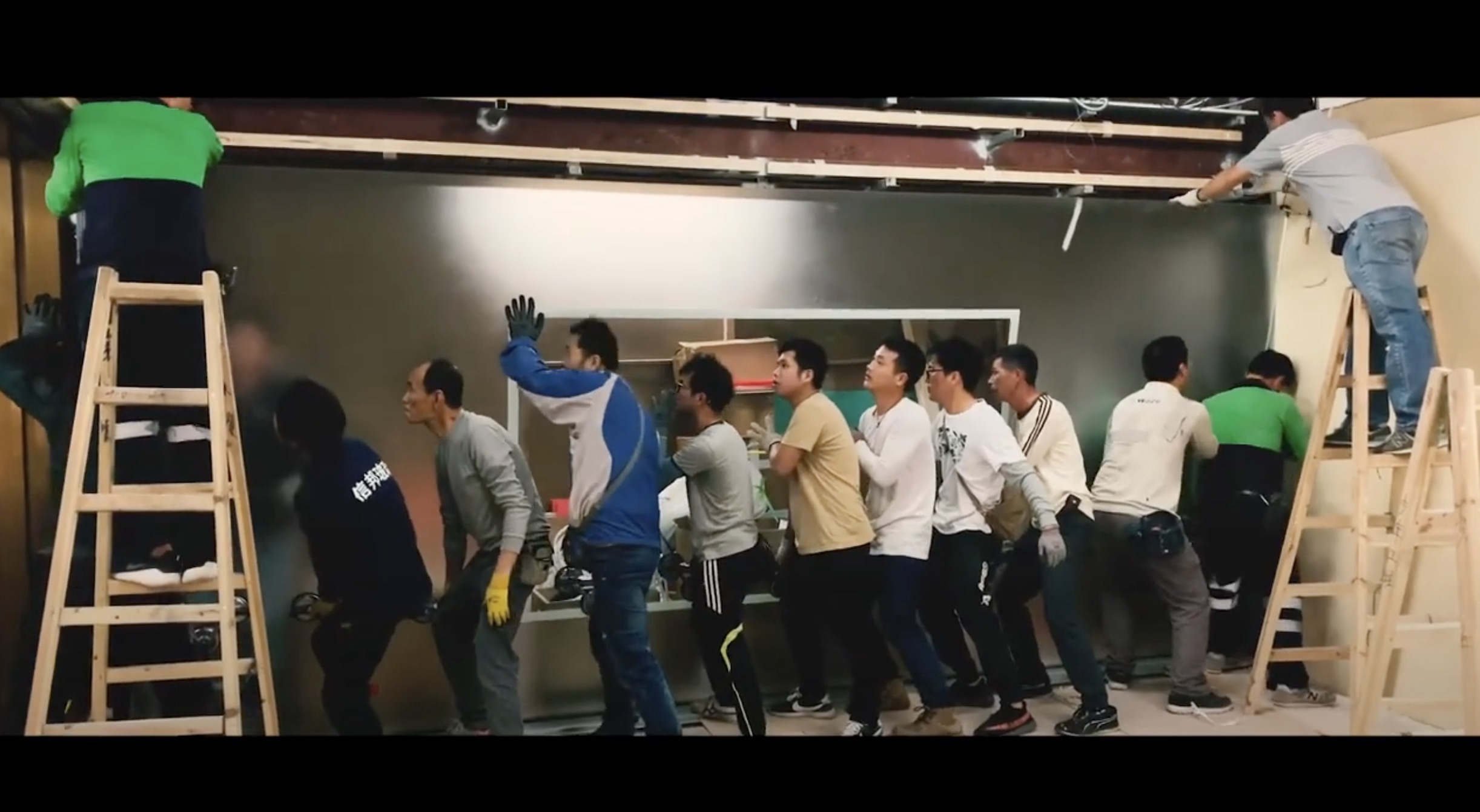 A group of people stand in a line, pushing against a wall, while two individuals on ladders work above them. The scene appears to be indoors, possibly during construction or renovation work.