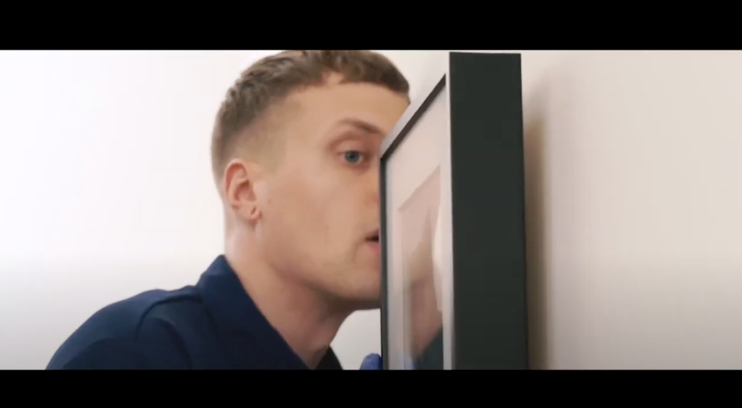 A person with short hair presses their face closely against the glass of a framed picture hanging on a white wall, looking intently at it.