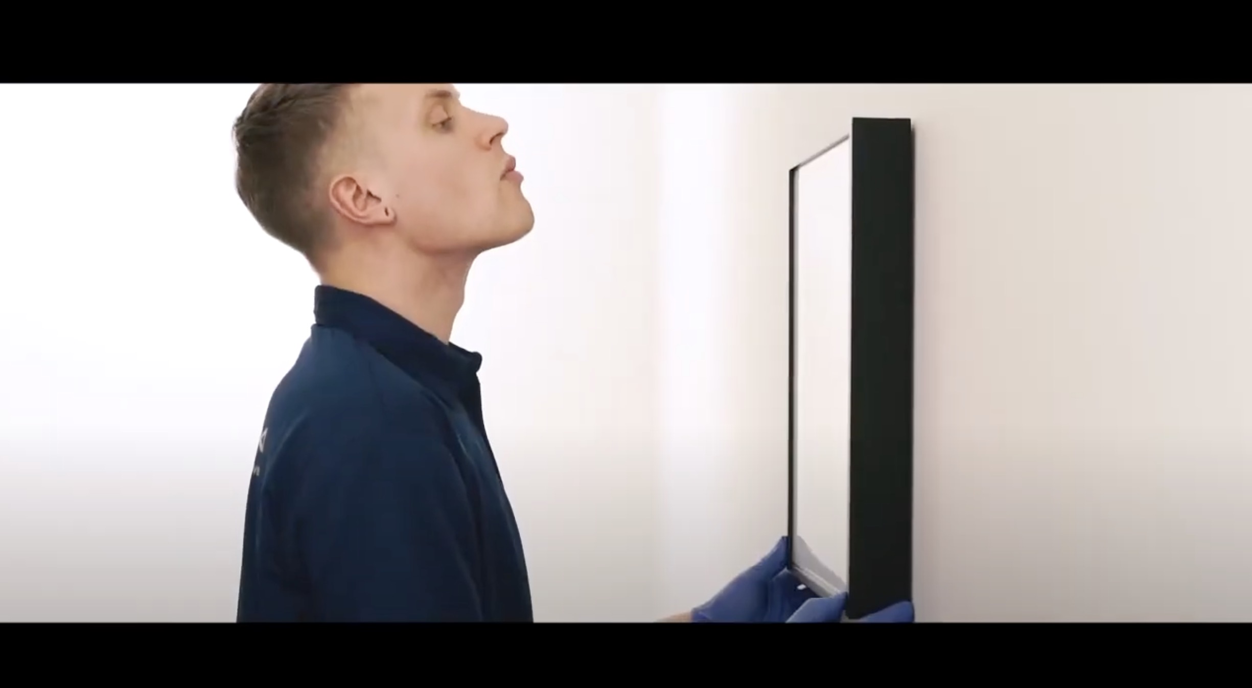 A person wearing blue gloves and a dark blue shirt carefully hangs a rectangular framed picture or mirror on a plain white wall, looking closely to ensure it is straight.