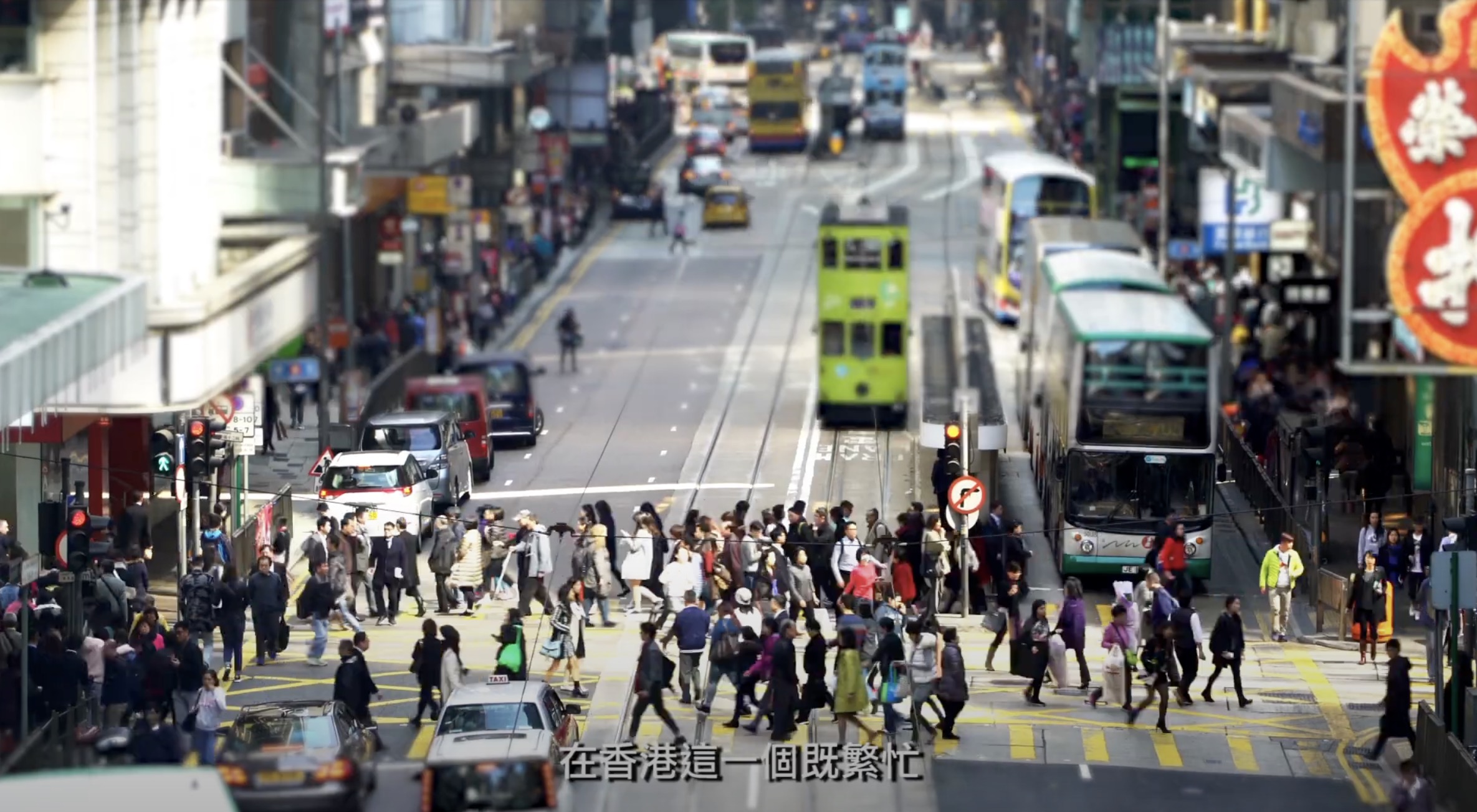 A busy street in Hong Kong with crowds crossing at a crosswalk, double-decker trams and buses on the road, and shops with signs. Chinese text appears at the bottom of the image.