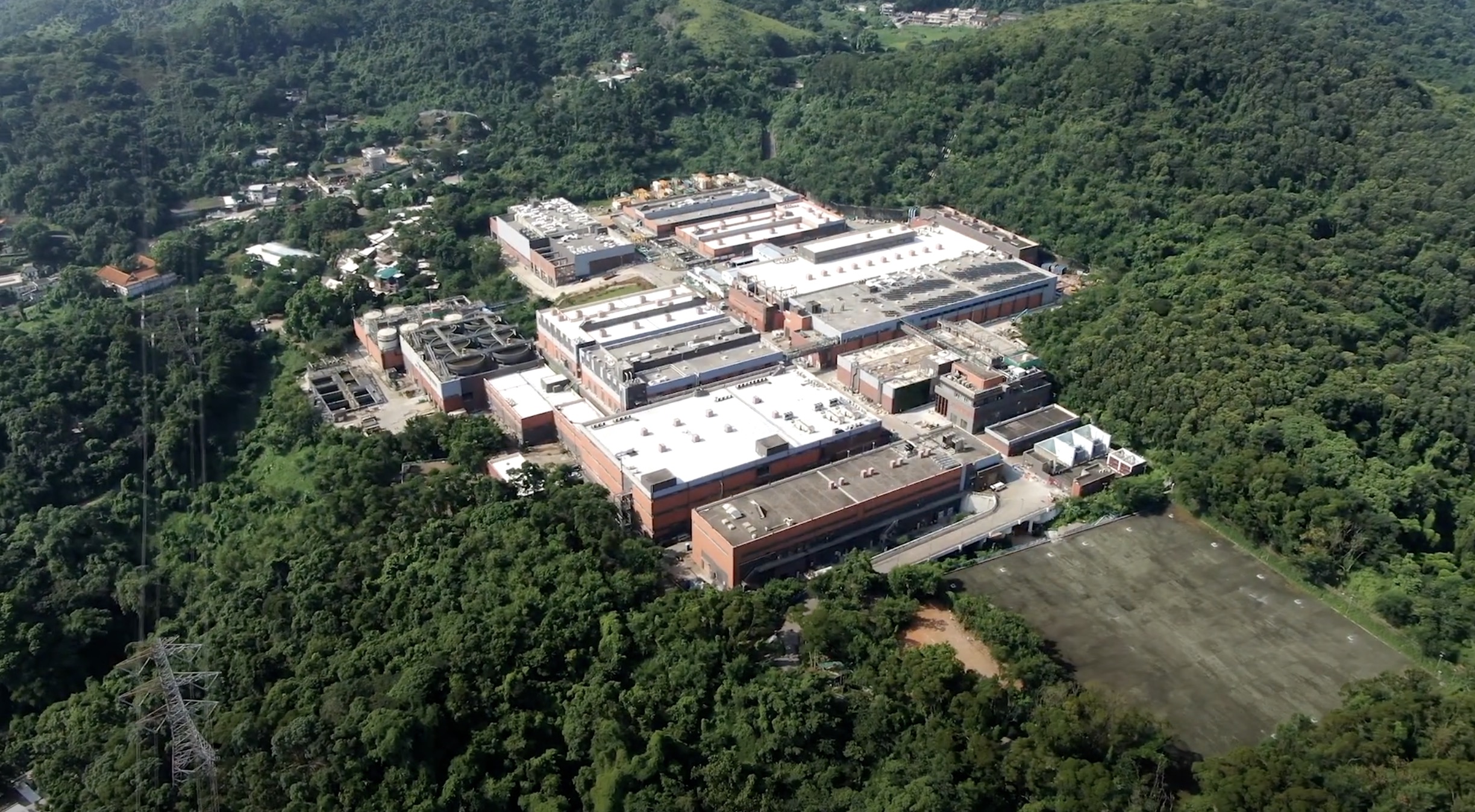 Aerial view of a large, rectangular building complex with red and white rooftops surrounded by dense green forest and hills, with some smaller structures and a cleared area visible nearby.