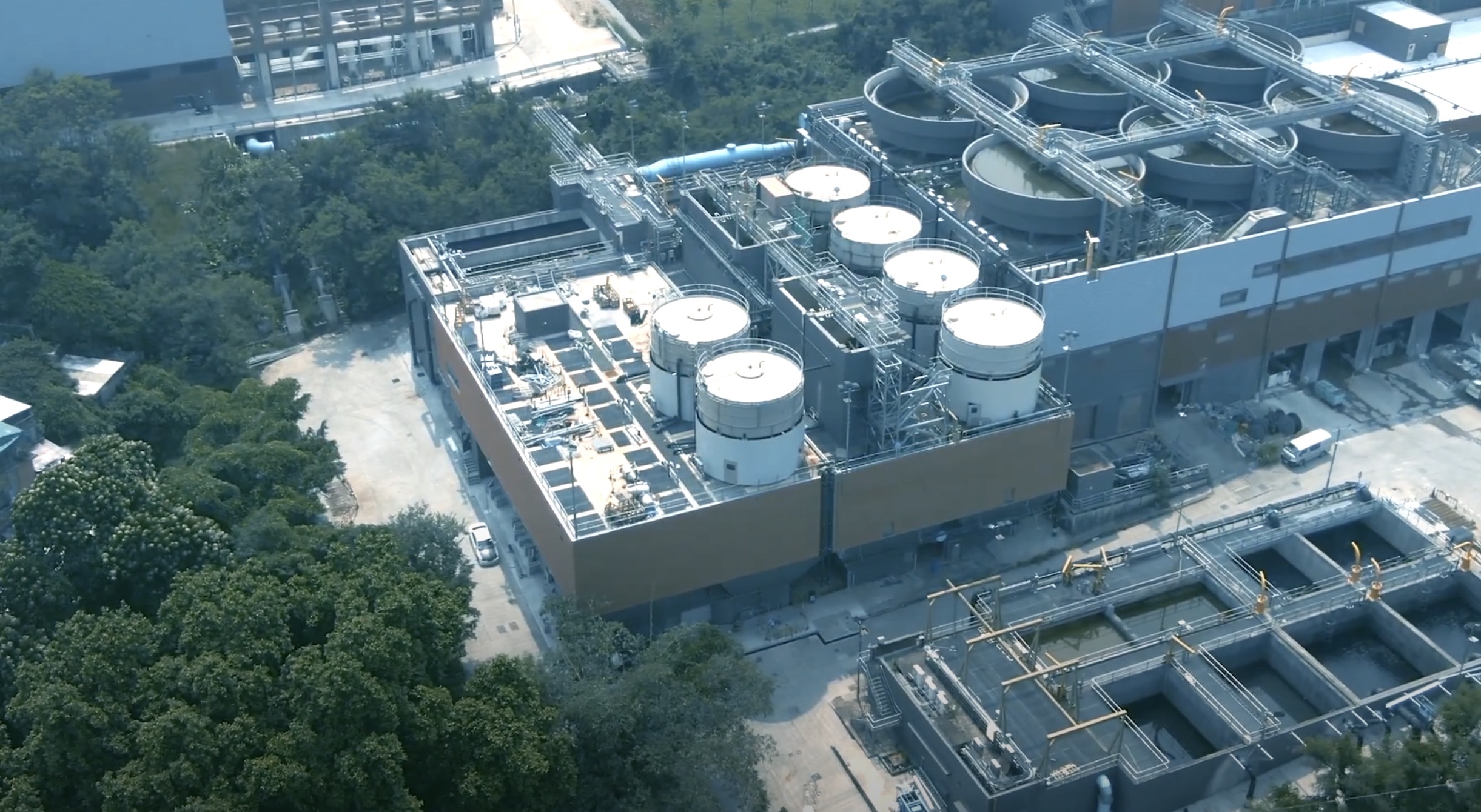Aerial view of an industrial facility with large cylindrical tanks, cooling towers, and complex piping surrounded by trees and adjacent structures.