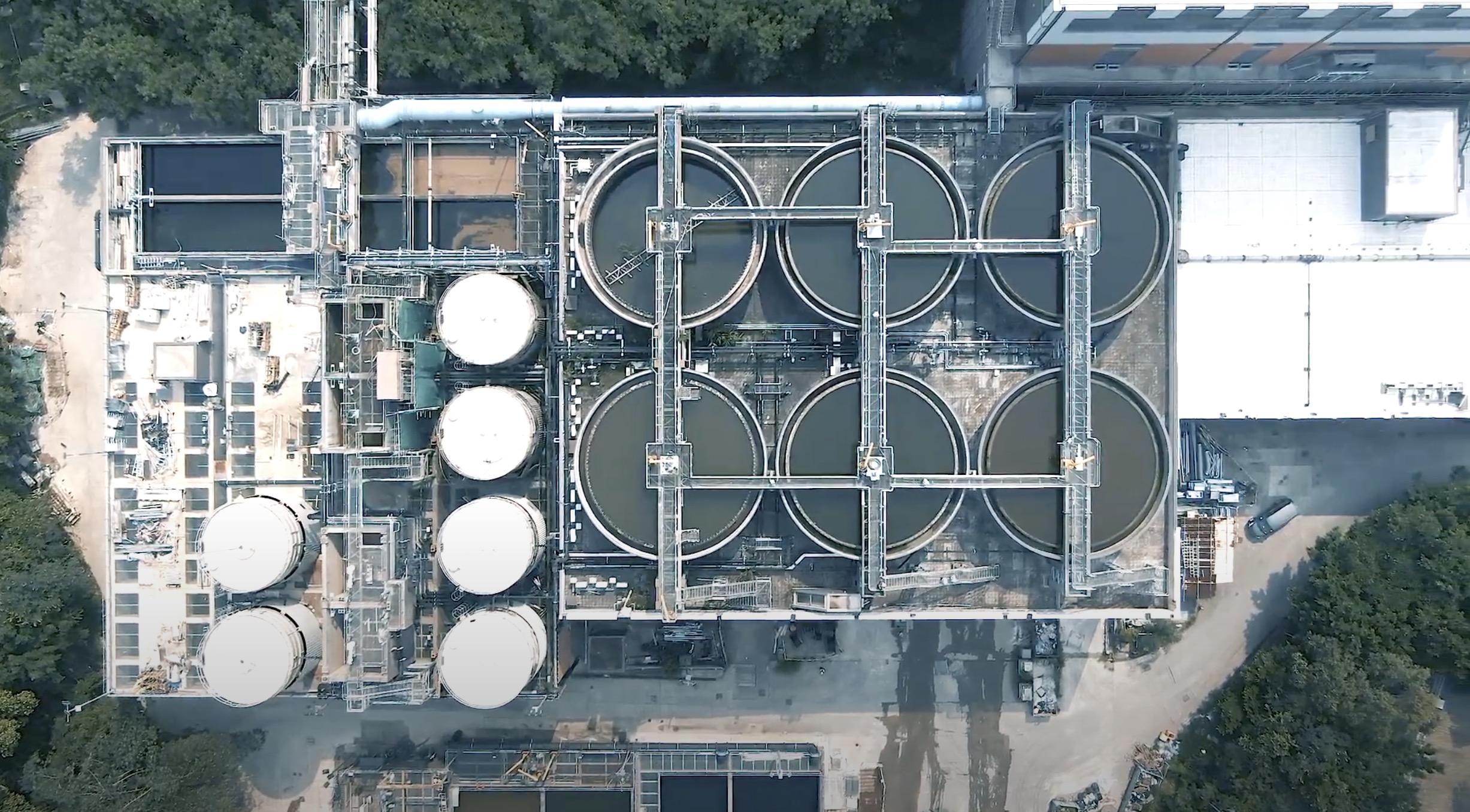 Aerial view of an industrial facility with six large circular tanks in two rows, four white cylindrical tanks on the left, and various pipes and structures; trees border the top edge of the site.