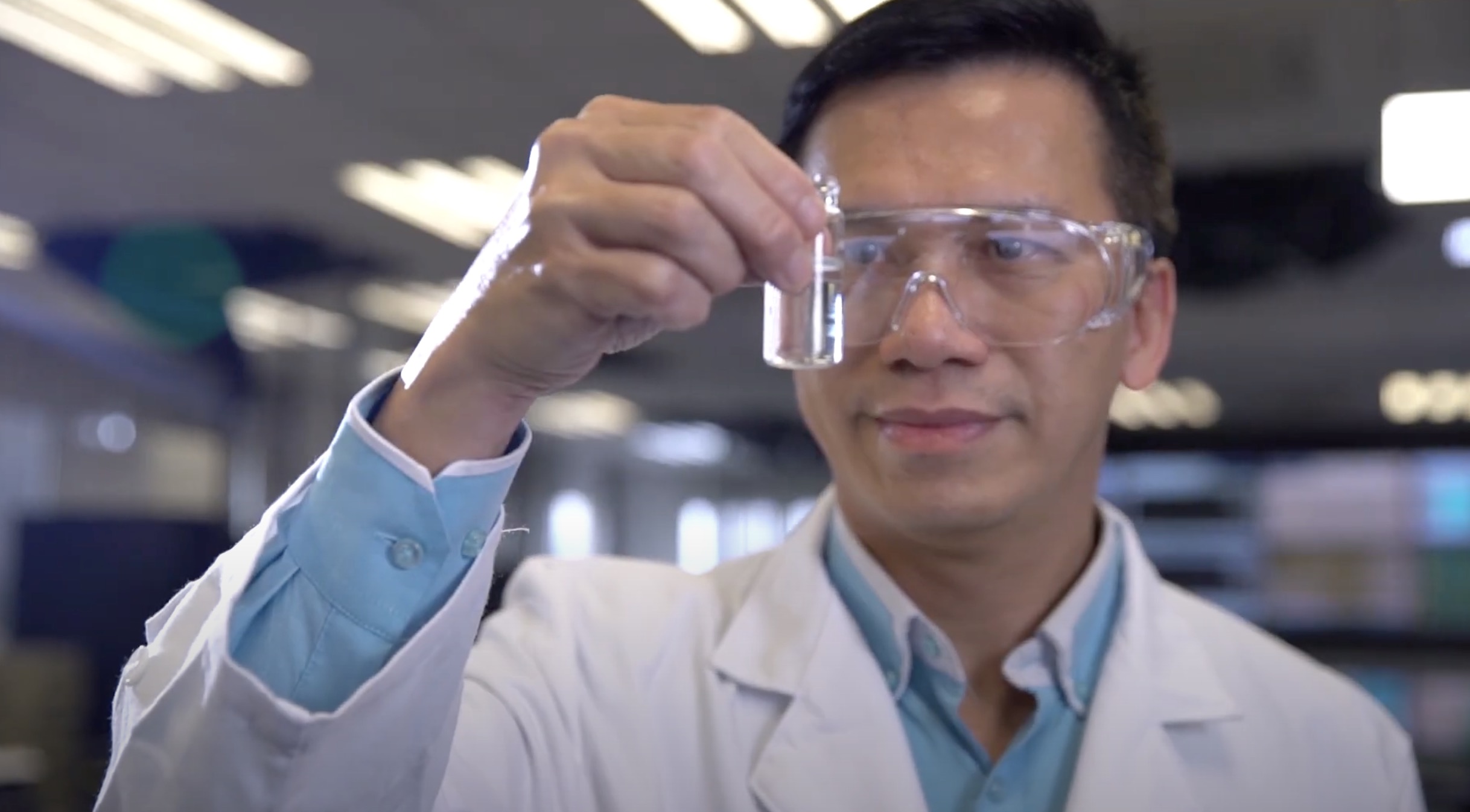A scientist in a white lab coat and safety goggles examines a clear liquid in a beaker, holding it up at eye level in a laboratory setting.