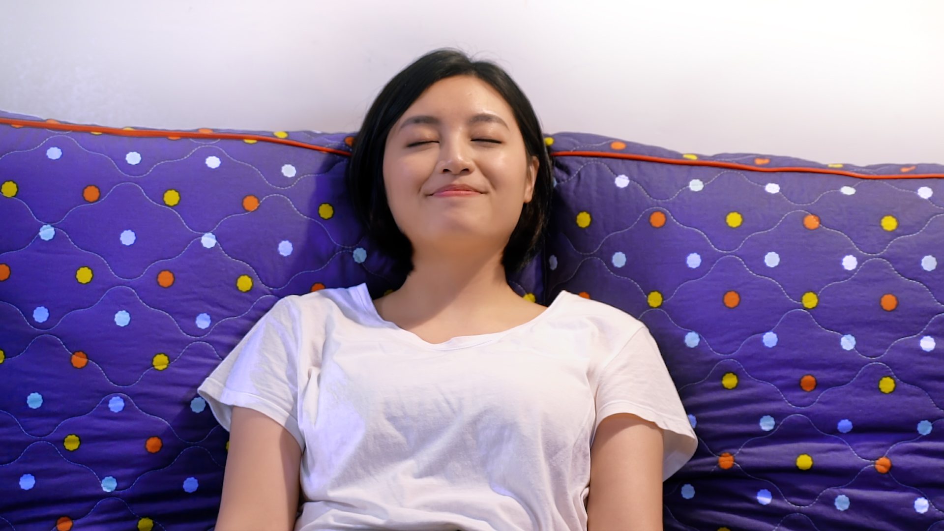 A woman with short black hair is lying on a bed with a blue and purple polka-dotted pillow, eyes closed, and smiling gently while wearing a white t-shirt.