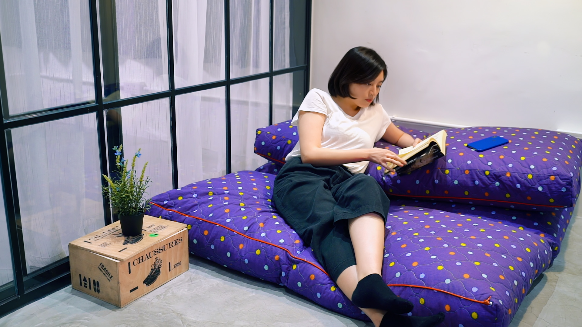 A woman sits on polka-dotted floor cushions by a large window, reading a book. A small potted plant and a blue notebook rest on a wooden crate nearby. The room has a relaxed and cozy atmosphere.