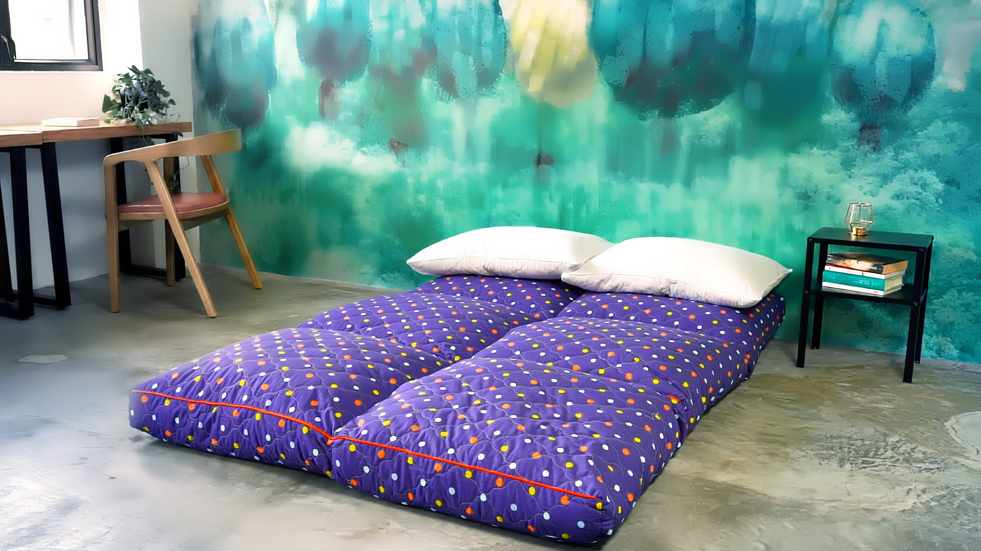 Two blue futon mattresses with small colorful dots are placed side by side on a concrete floor, topped with white pillows. Behind them is a green and blue abstract wall, with a small table and chair nearby.