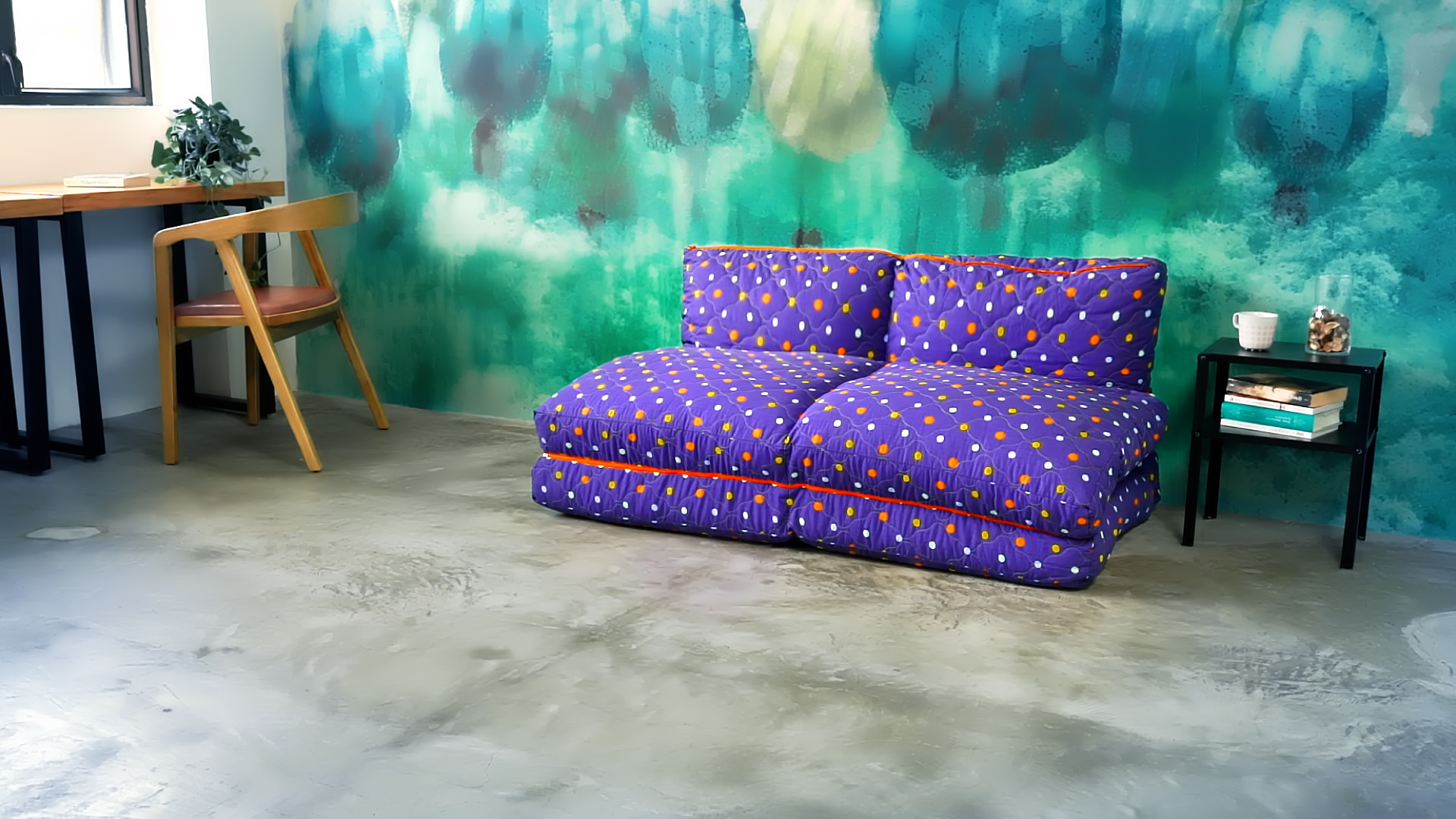 A cozy room with a purple patterned floor sofa, a green and blue abstract wall, a small black side table with books and a jar, and a wooden desk with a chair and a potted plant by the window.
