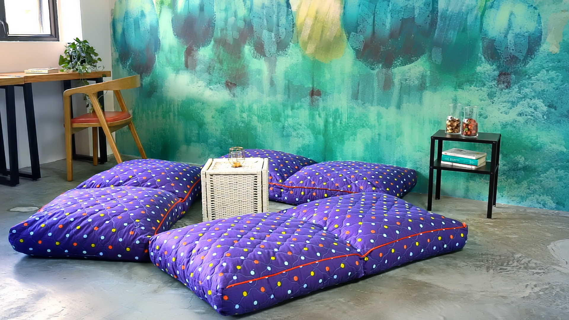 Four large purple floor cushions with colorful dots are arranged around a low white wicker table in a modern room with a green and blue abstract wall, a window, and a wooden desk with a chair and plant.
