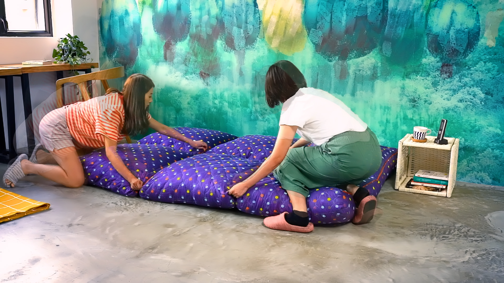 Two people are laying out a colorful purple comforter with small patterns on the floor of a room with a vibrant teal and yellow painted wall and a small table holding books and a cup.
