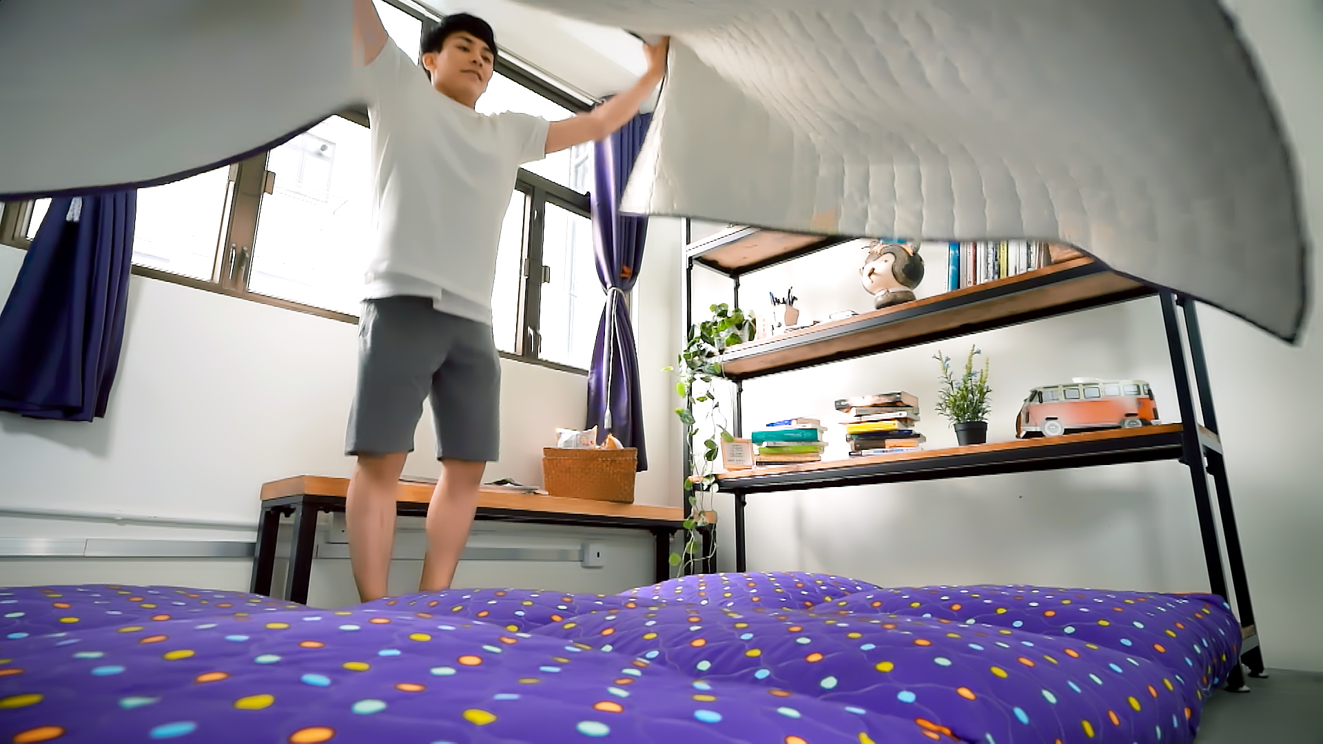 A person in a white t-shirt and gray shorts spreads a blanket or comforter over a colorful, polka-dotted bed in a bright bedroom with bookshelves, plants, and large windows.