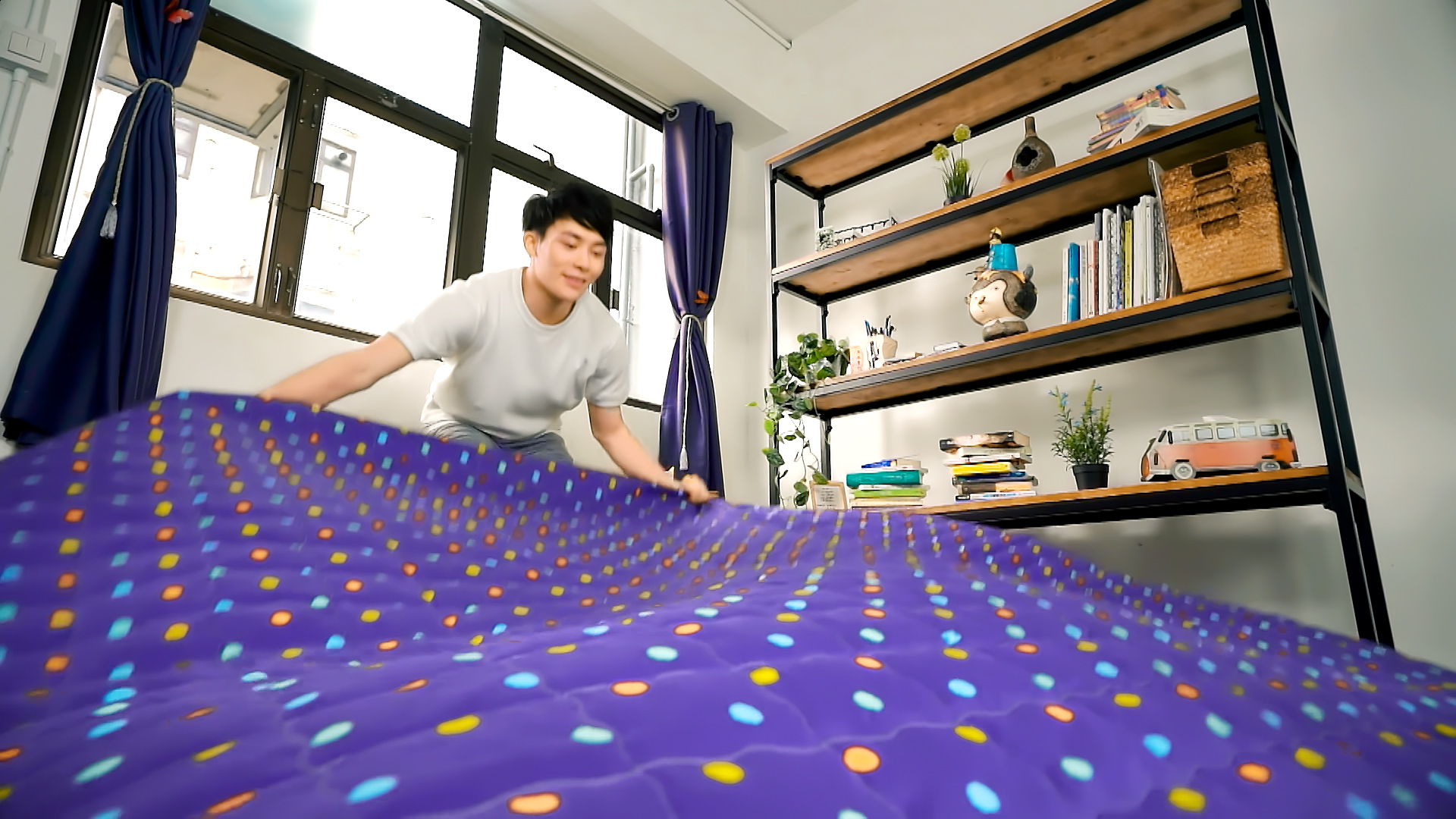 A person in a white shirt is spreading a purple bedsheet with colorful dots over a bed in a bright, organized room with bookshelves, plants, and large windows.