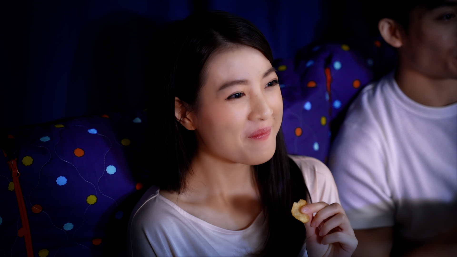 A young woman with long dark hair smiles while eating a snack, sitting in a seat with a colorful polka dot pattern, possibly in a movie theater.