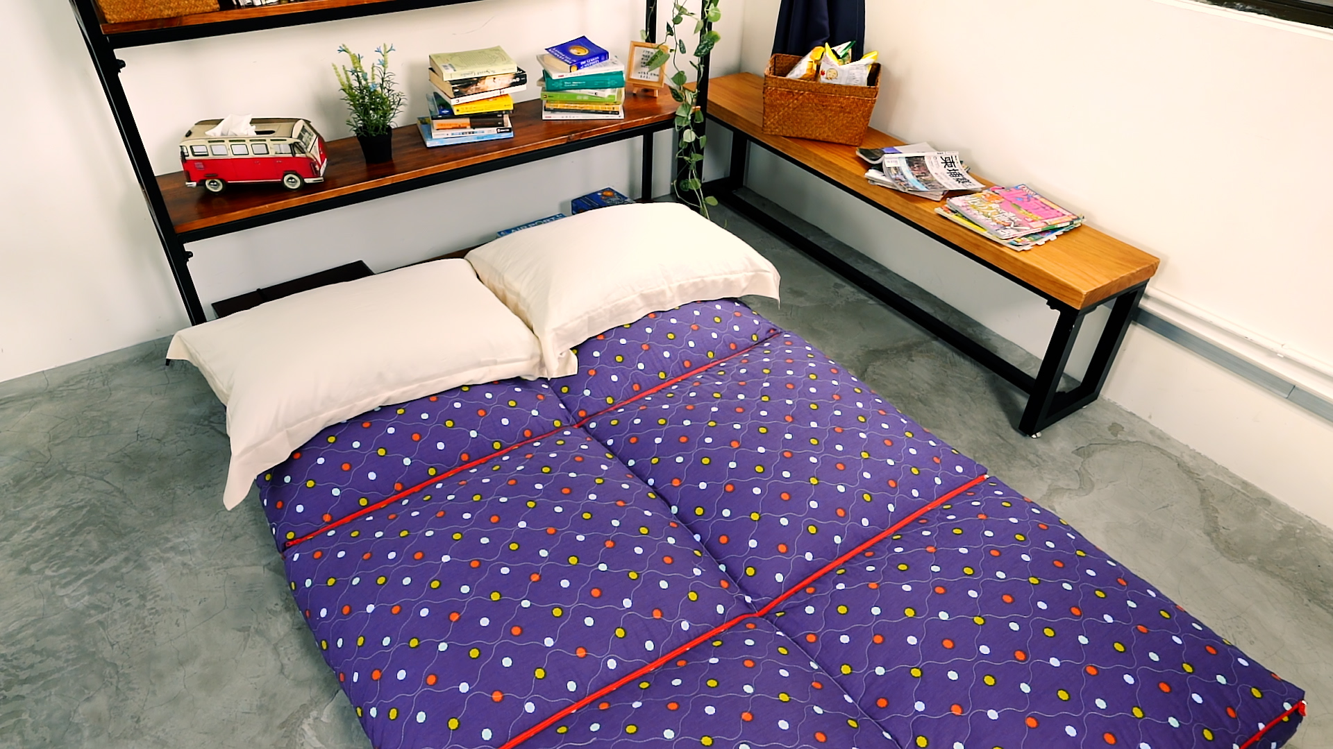 A futon with a purple, polka-dotted cover and two pillows is laid out on a concrete floor next to a wooden shelf with books, plants, and a toy van, and a bench with magazines in a bright, cozy room.