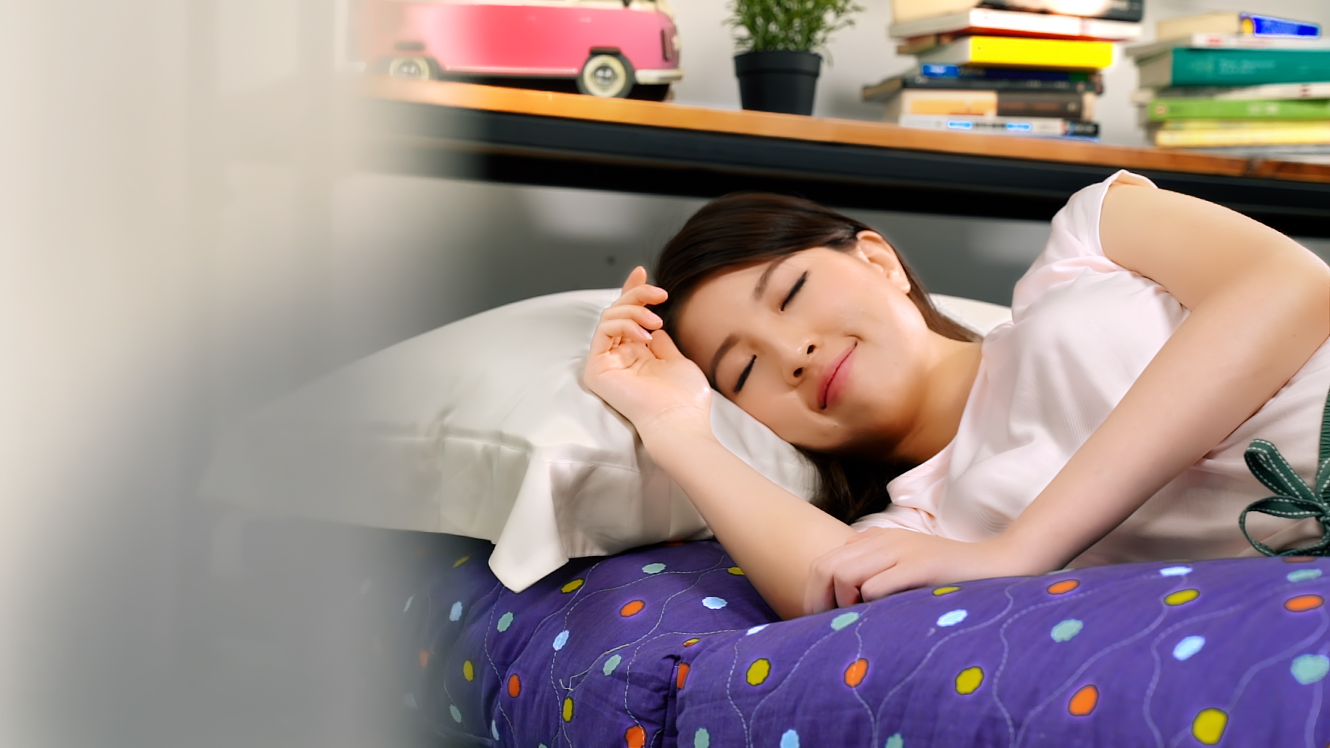 A young woman with long dark hair sleeps peacefully on a bed with a blue polka-dotted blanket. She is smiling slightly, and there are books, a plant, and a pink toy van on a shelf in the background.