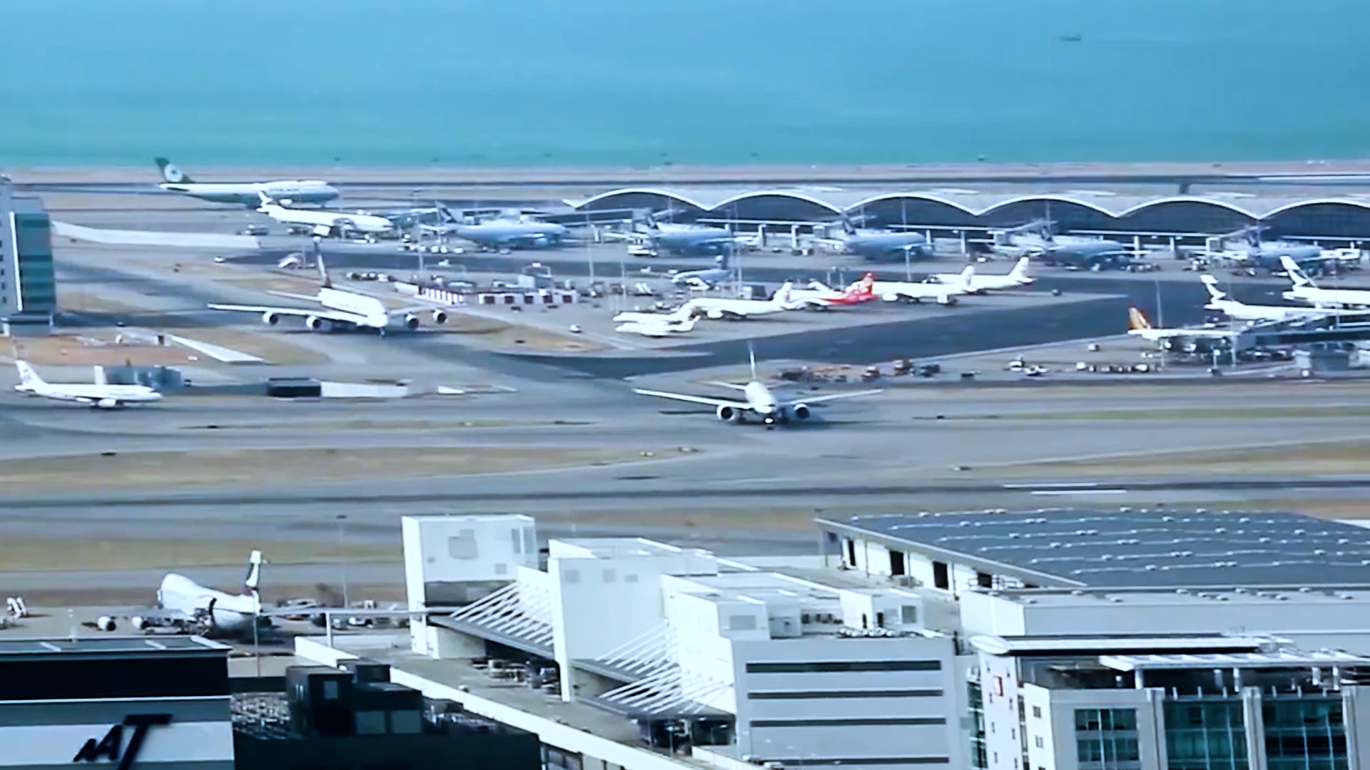 A busy airport scene with multiple airplanes on runways and parked near terminals; the terminal building has a curved roof, and the ocean is visible in the background.