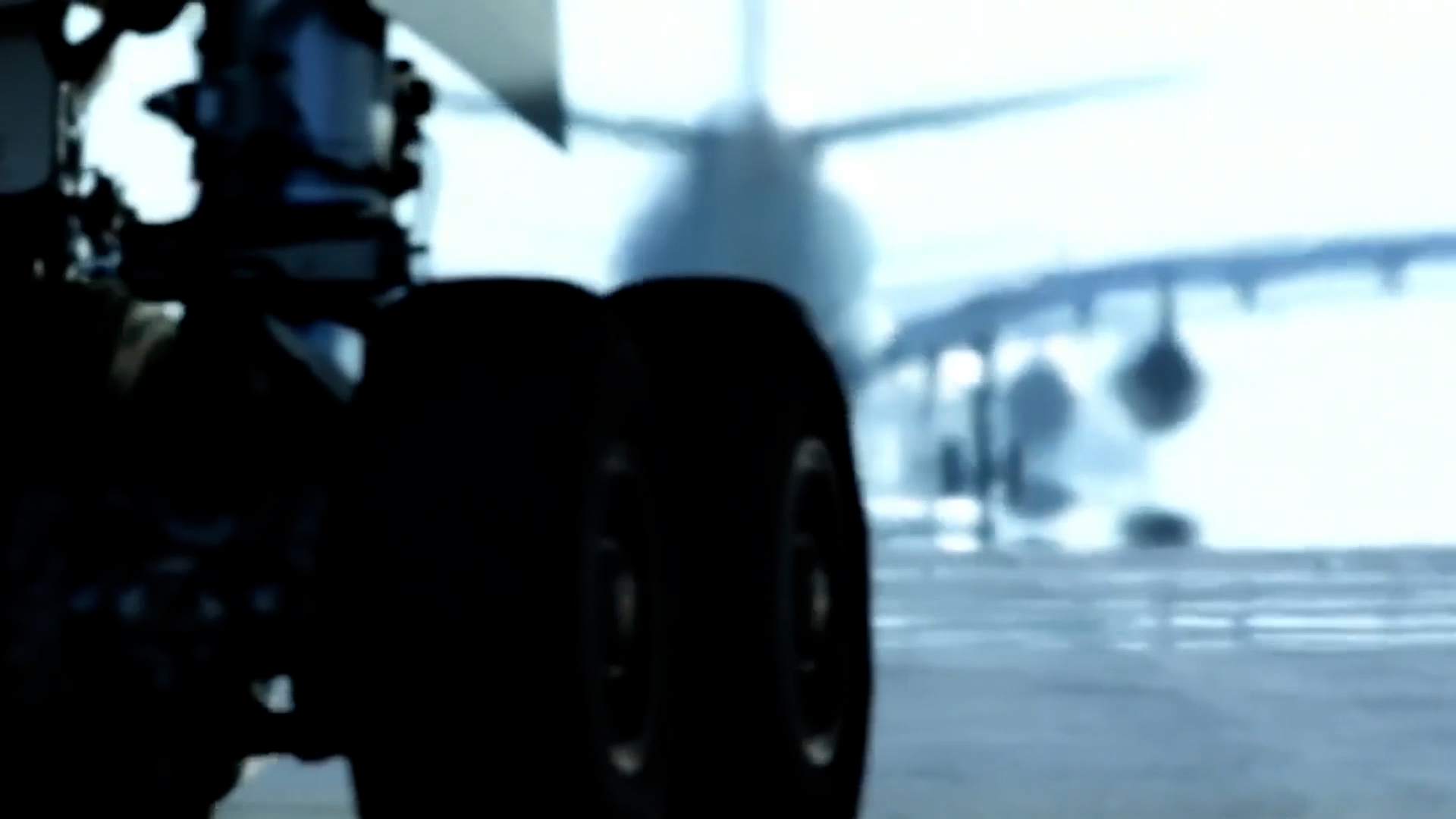 Close-up of airplane landing gear on a tarmac with a blurred background showing another airplane and airport structures in bright daylight.