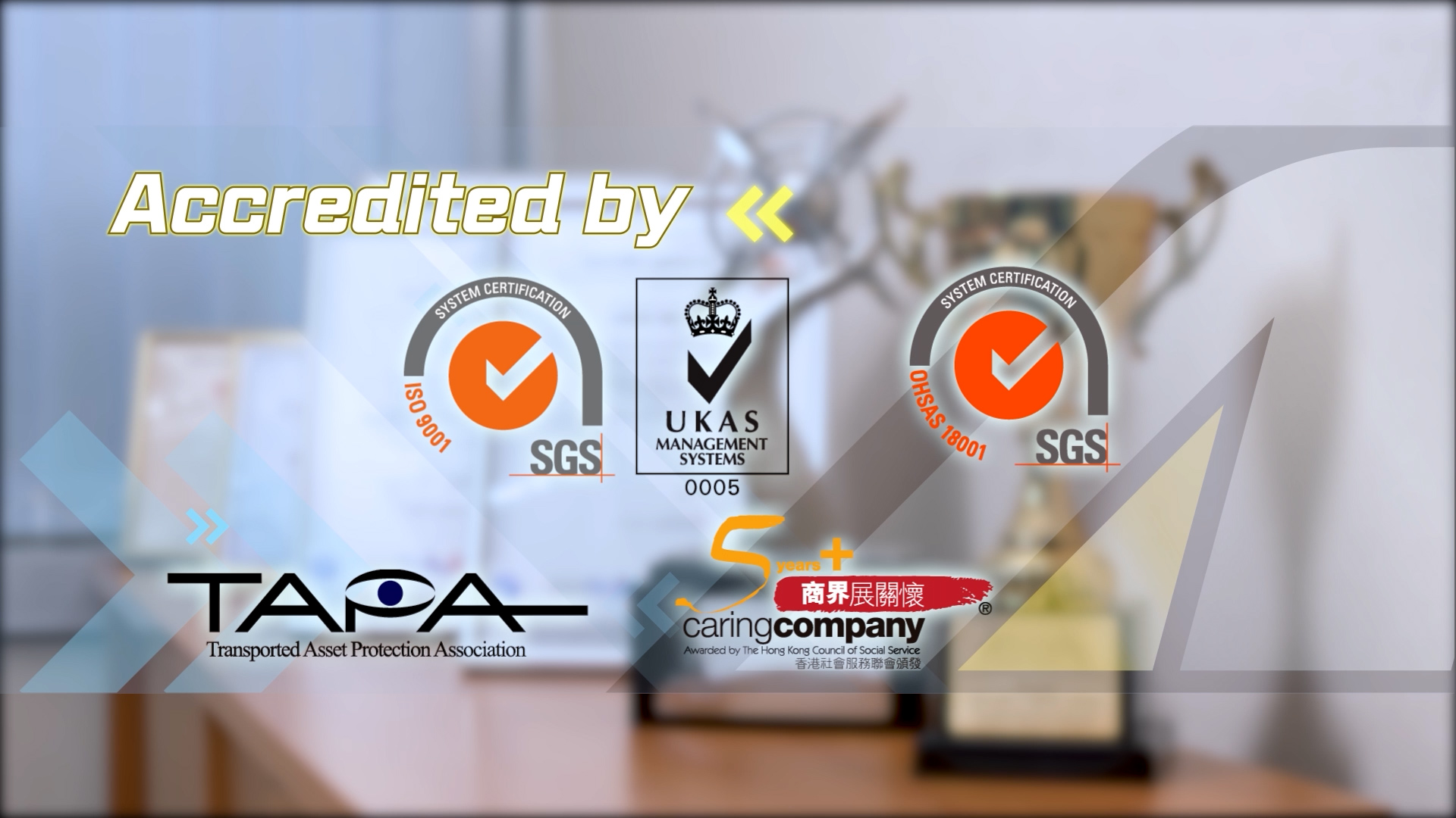 Glass panel displaying certification logos, including ISO 9001, OHSAS 18001, SGS, UKAS, TAPA, and Caring Company, with a blurred trophy and office background. Text reads "Accredited by.
