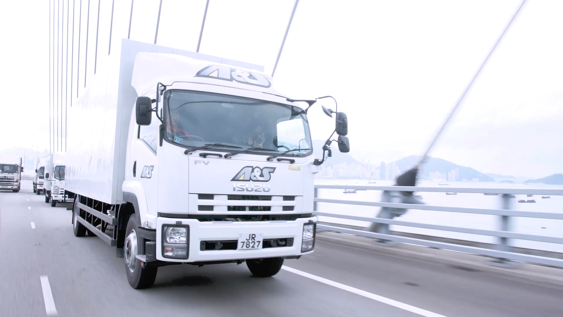 A white Isuzu delivery truck with "A&S" branding drives on a bridge, with another truck visible behind it. The background shows a city skyline and water on a cloudy day.