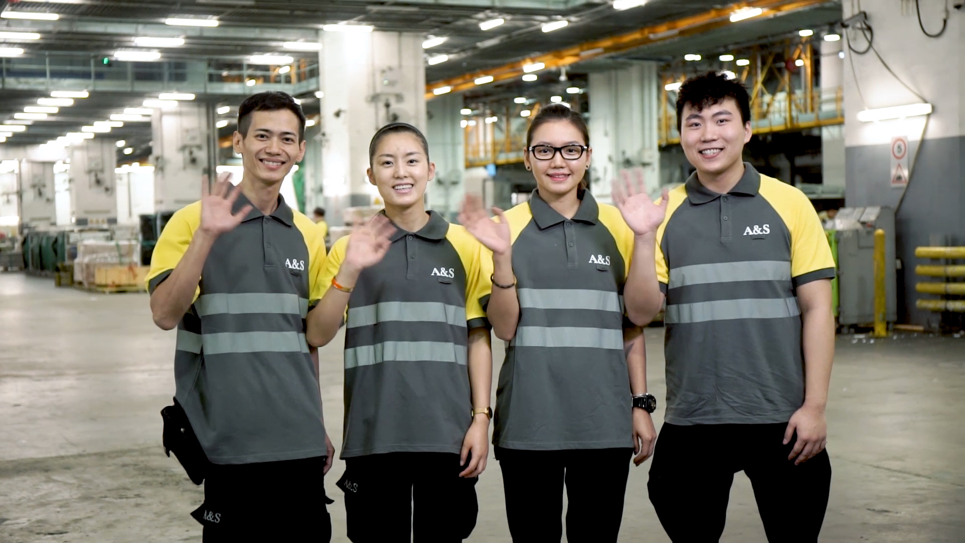 Four people in matching gray and yellow uniforms stand indoors, smiling and waving at the camera. The setting appears to be an industrial or warehouse facility with bright lighting and equipment in the background.