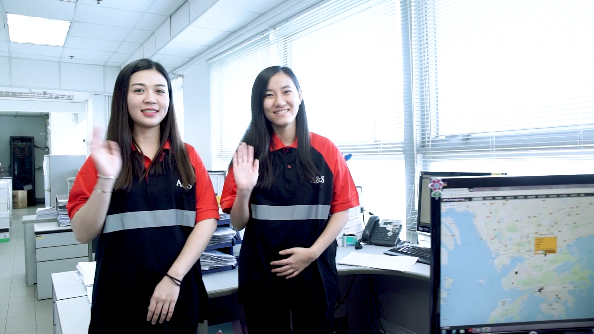 Two women wearing matching black and red uniforms stand in an office, smiling and waving at the camera. Behind them are desks, office equipment, and large windows. A computer screen with a map is visible in the foreground.