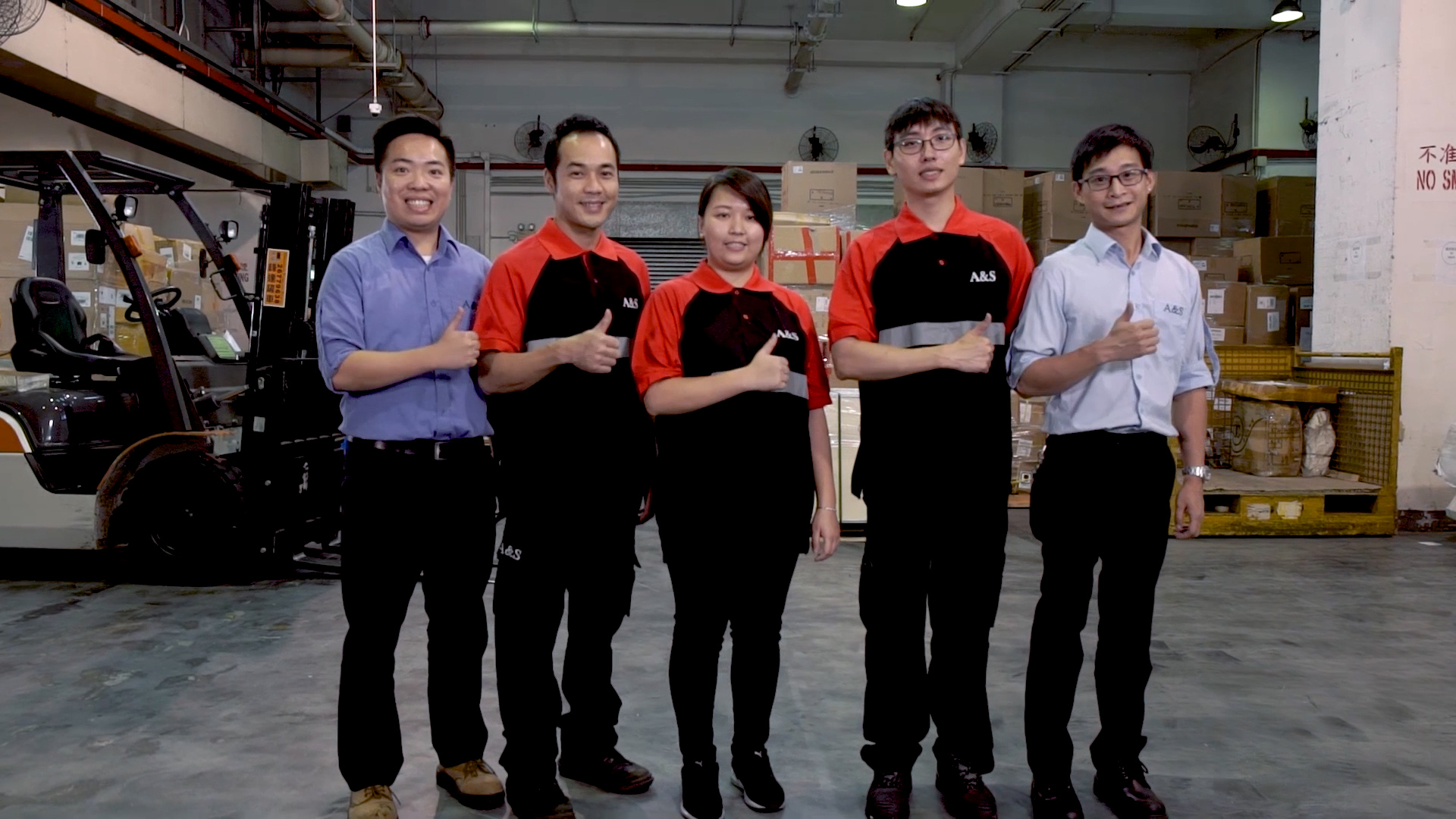 Six people stand indoors in a warehouse, posing for a group photo. Three wear black-and-red uniforms, while the others wear business attire. They all smile and give a thumbs-up gesture, with stacked boxes and equipment in the background.