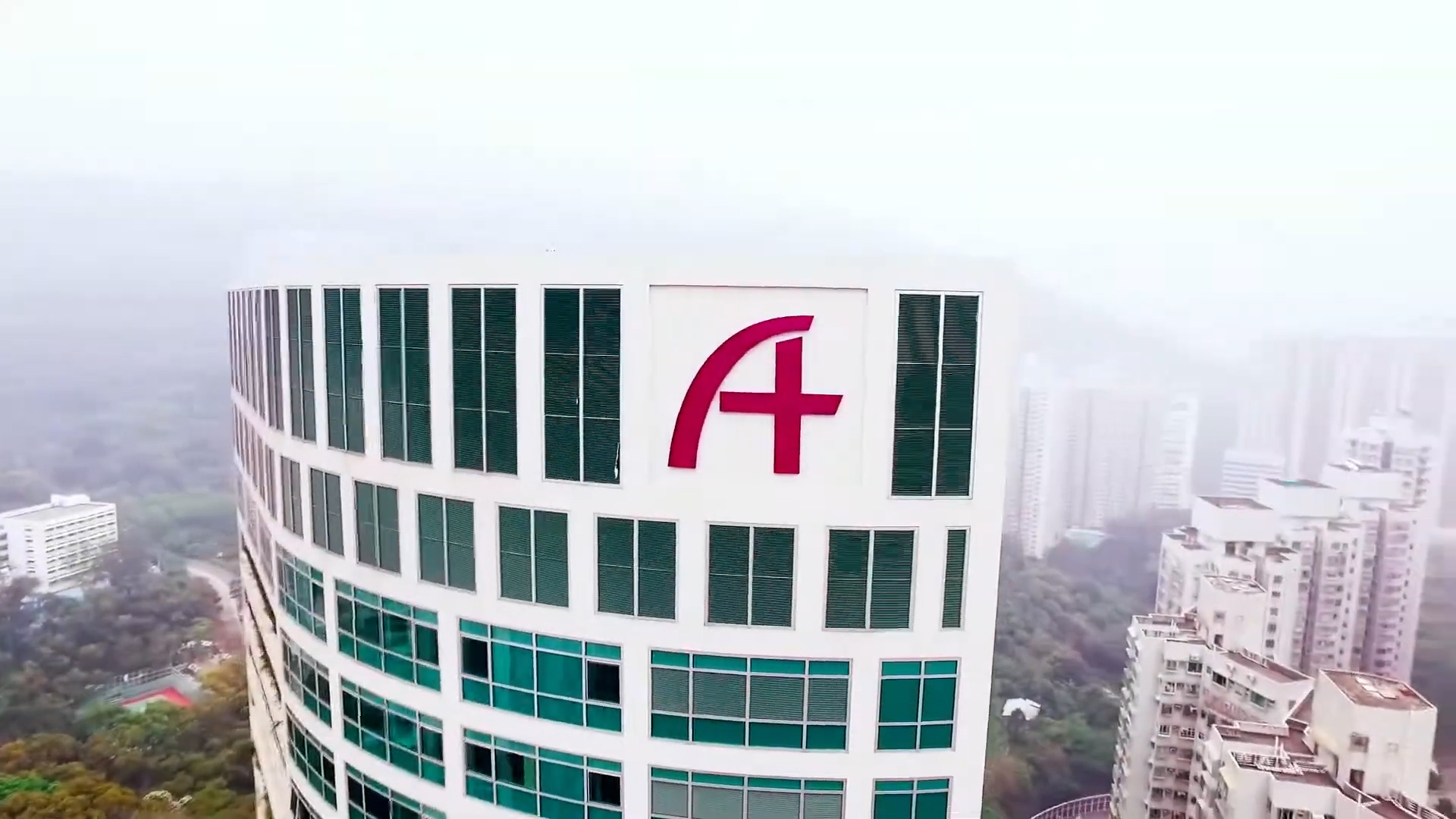 A tall, white, modern building with many green-tinted windows features a large red letter "A" logo on its upper facade. High-rise buildings and a foggy cityscape are visible in the background.