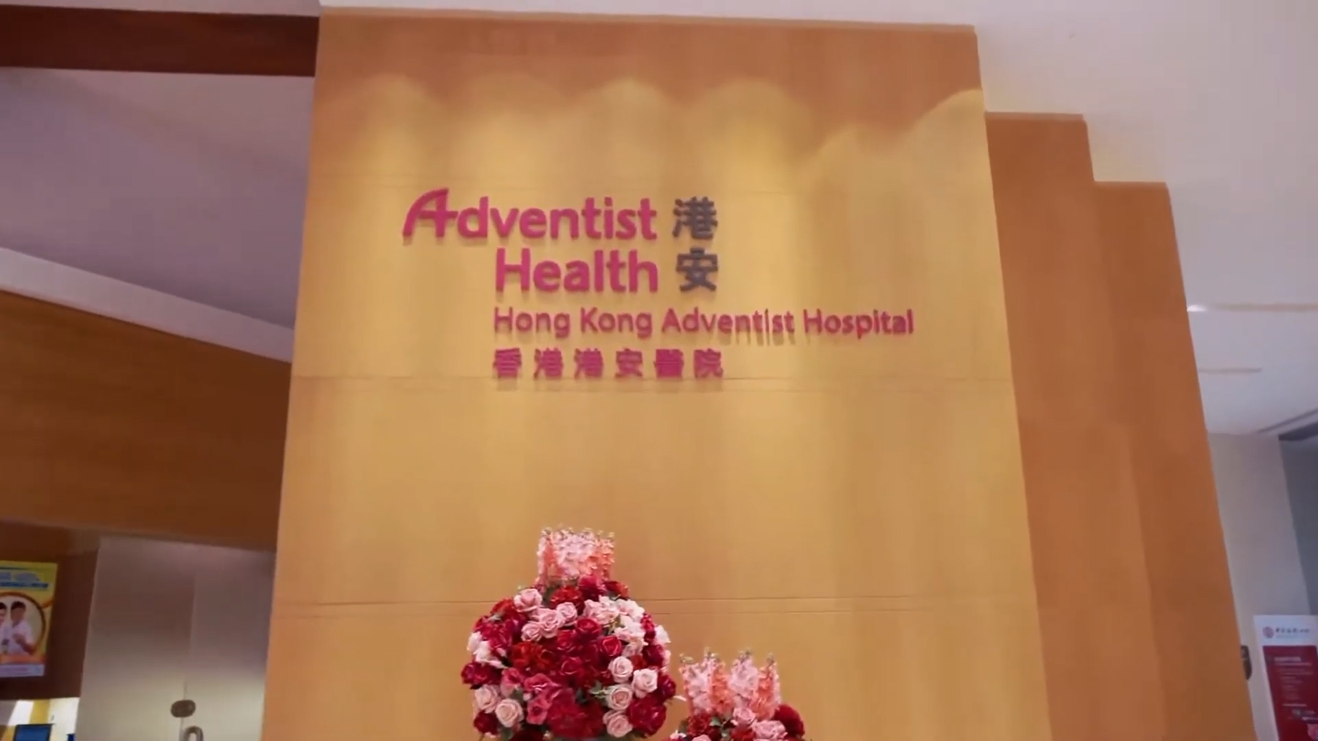A wall with the Hong Kong Adventist Hospital logo in English and Chinese, with a bouquet of red and white flowers arranged in front of it.
