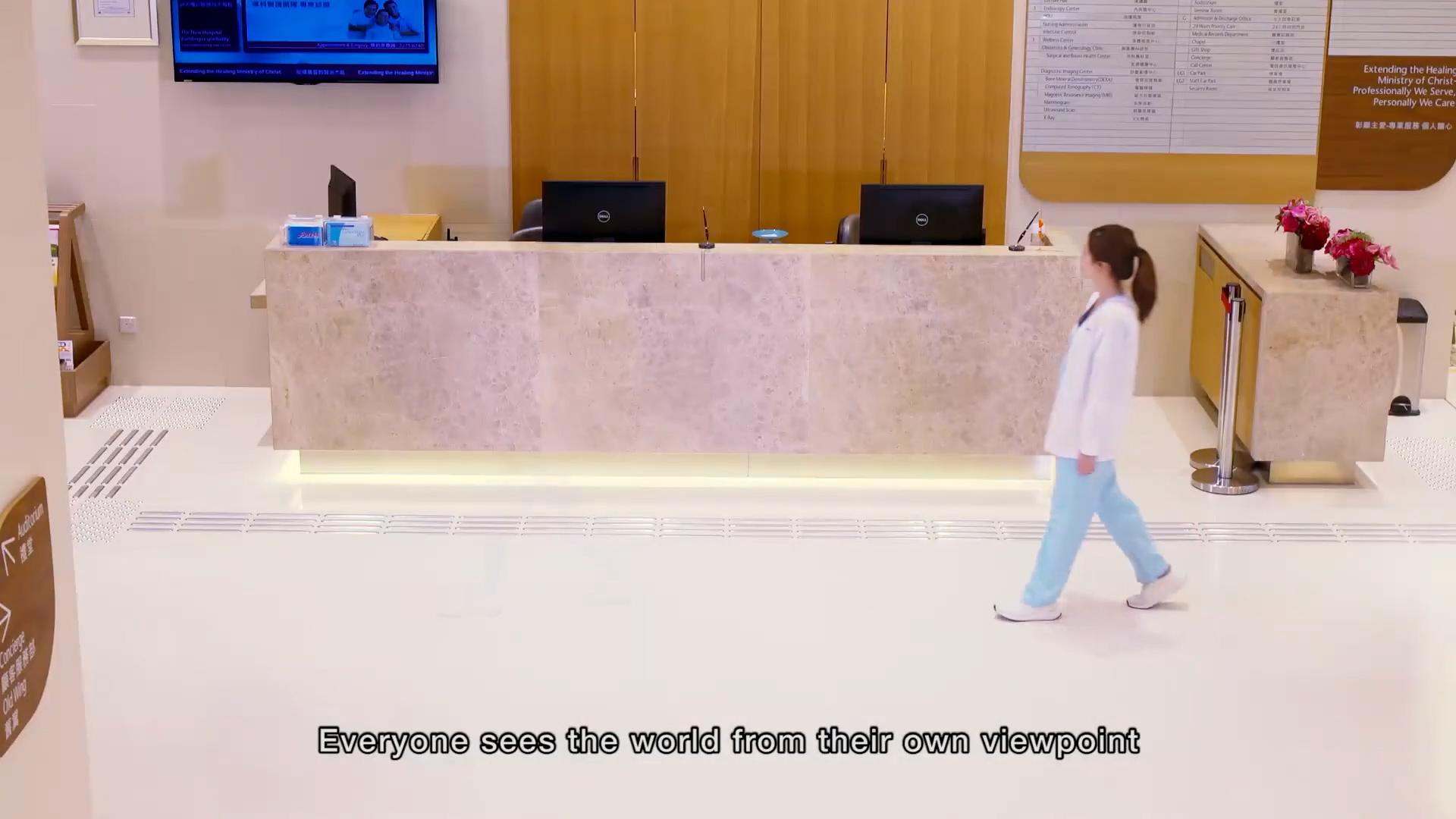 A person in medical scrubs walks past a modern, marble-fronted reception desk in a bright lobby. A TV and large sign are visible on the wall. Subtitle reads: "Everyone sees the world from their own viewpoint.