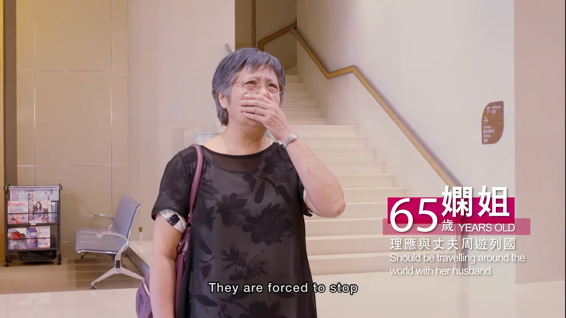 An older woman with short gray hair, glasses, and a black floral shirt stands near stairs, covering her mouth with her hand. Onscreen text notes she is 65, should be traveling with her husband, and subtitles read, "They are forced to stop.