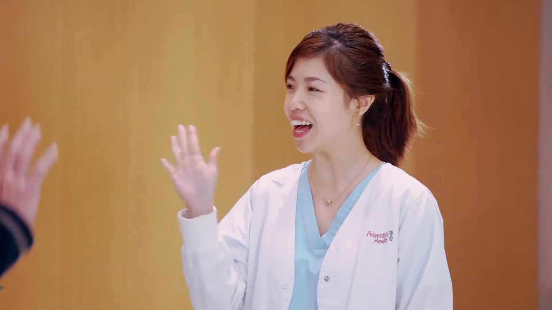 A woman in a white medical coat smiles and waves with her right hand. She has dark hair tied back and is standing against a plain, light-colored background. Another person's hand is visible in the foreground, also raised.