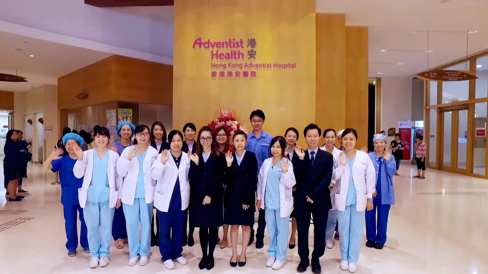 A group of medical staff and professionals, including doctors and nurses, stand together indoors at Hong Kong Adventist Hospital, smiling and waving at the camera in front of a hospital sign.