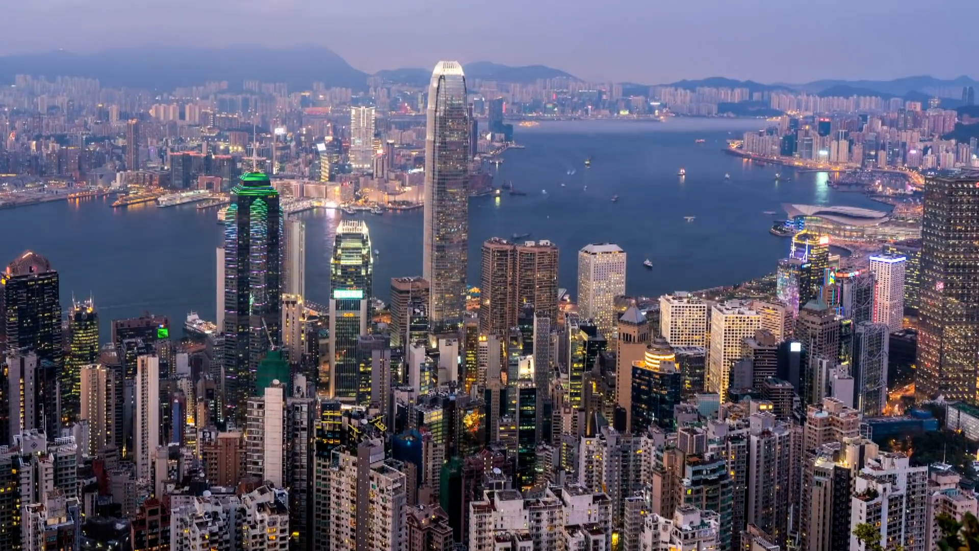 Aerial view of a dense city skyline at dusk with tall skyscrapers, illuminated buildings, and a harbor with boats, set against distant mountains under a soft evening sky.