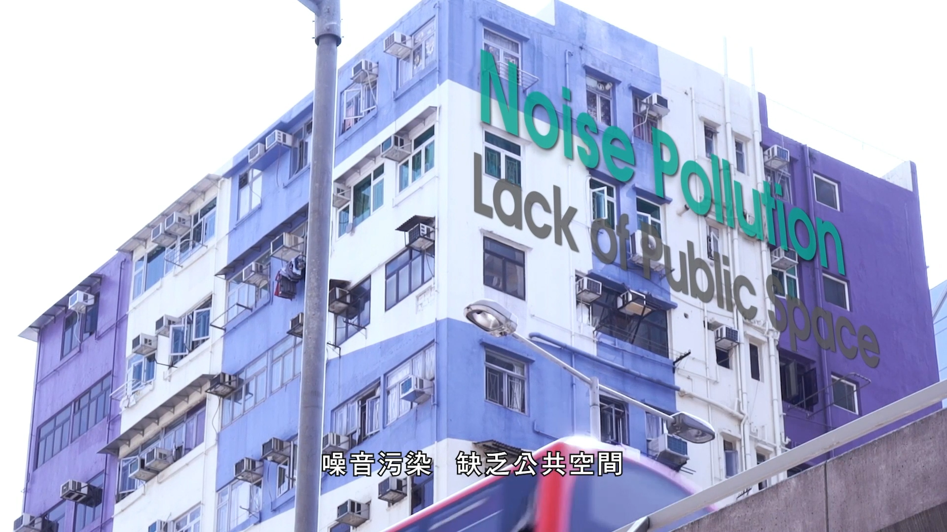 A blue and white apartment building with many air conditioners has large text reading "Noise Pollution Lack of Public Space." Chinese subtitles appear at the bottom of the image. A blurred object is visible in the foreground.