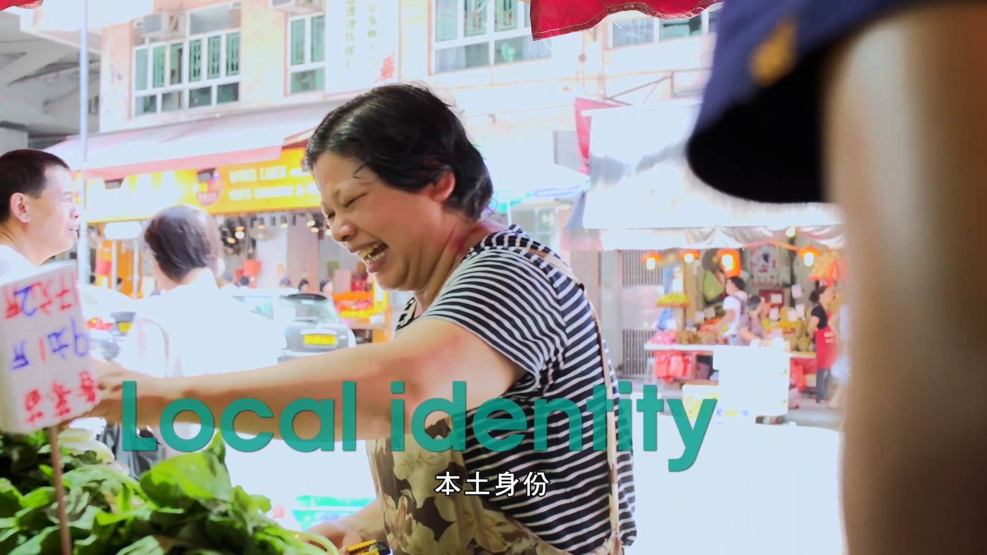 A woman in a striped shirt and apron smiles while selling vegetables at an outdoor market. The words "Local identity" and Chinese text are overlaid on the image. Bright shops and produce are visible in the background.