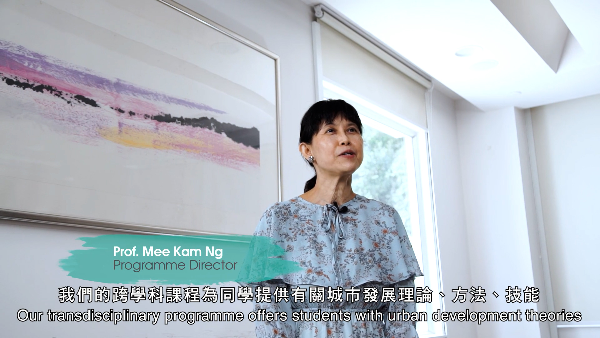 A woman stands and speaks in a bright room with a framed abstract painting on the wall. Text on the image introduces her as Prof. Mee Kam Ng, Programme Director, with subtitles about an urban development programme.