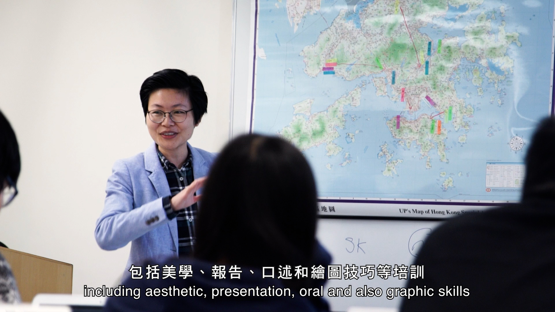 A person in glasses and a blue blazer speaks and gestures in front of a map of Hong Kong, with subtitles in Chinese and English about training in aesthetic, presentation, oral, and graphic skills.
