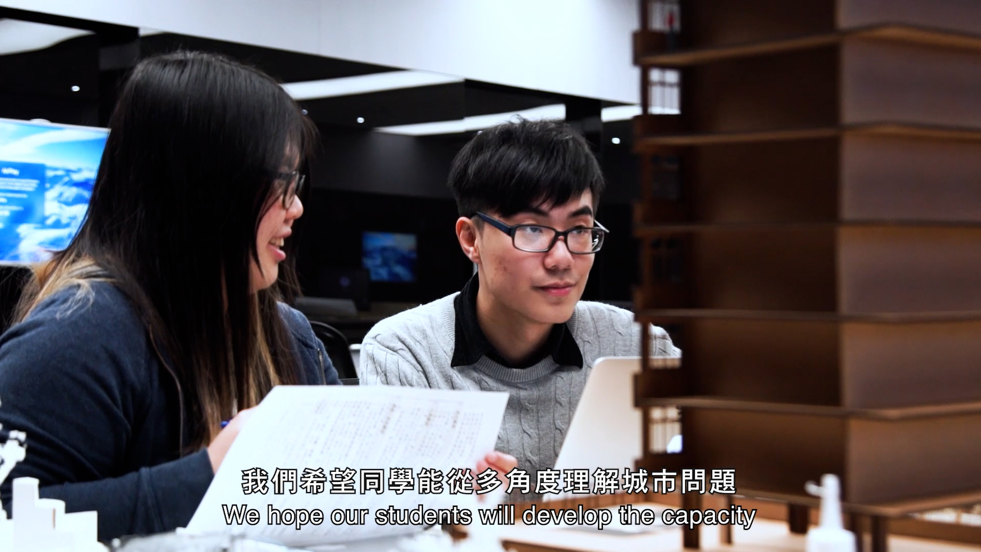 Two students sit together at a desk with papers, smiling and looking at something off-screen. In the foreground is a wooden architectural model. Subtitles in Chinese and English are visible at the bottom.