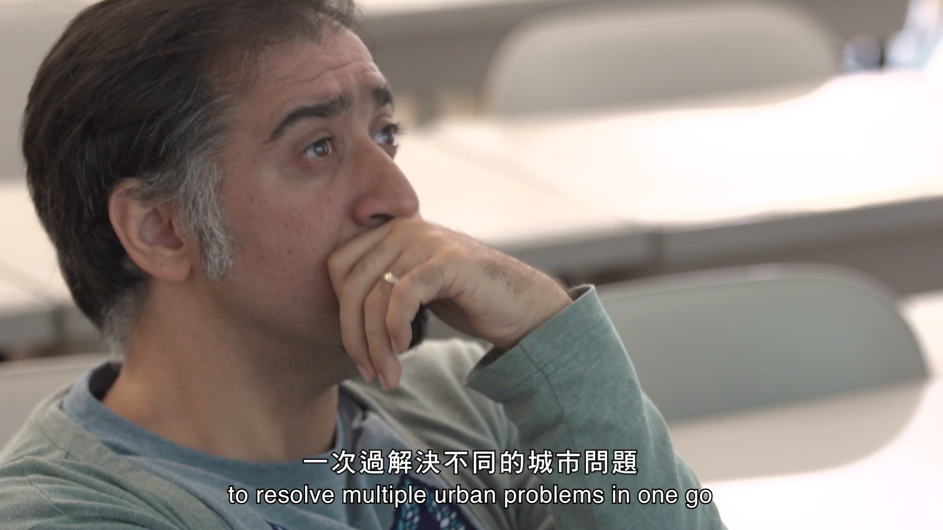 A man with dark hair and a gray shirt sits thoughtfully with his hand on his chin in a classroom. Chinese and English subtitles at the bottom read: "to resolve multiple urban problems in one go.