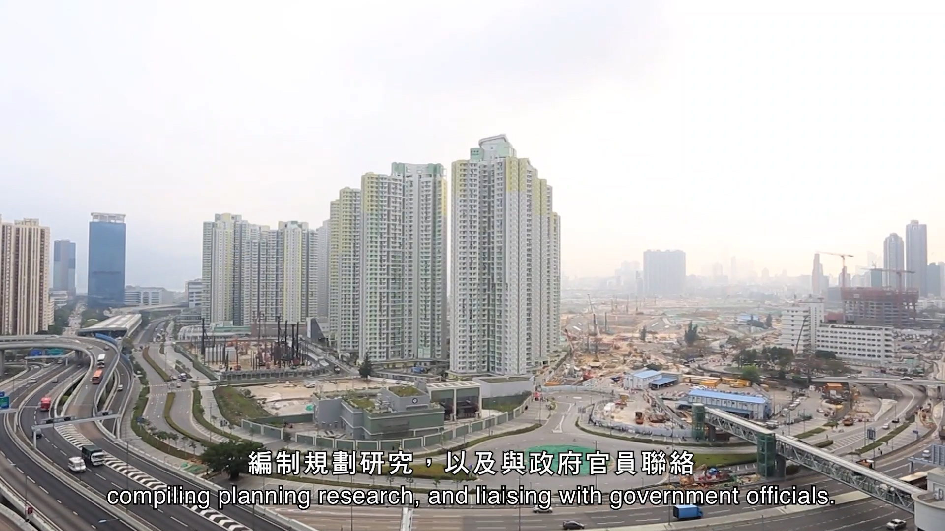 A cityscape featuring tall residential buildings, busy roads, and a construction site, with hazy sky in the background. Subtitles at the bottom read: “compiling planning research, and liaising with government officials.”.