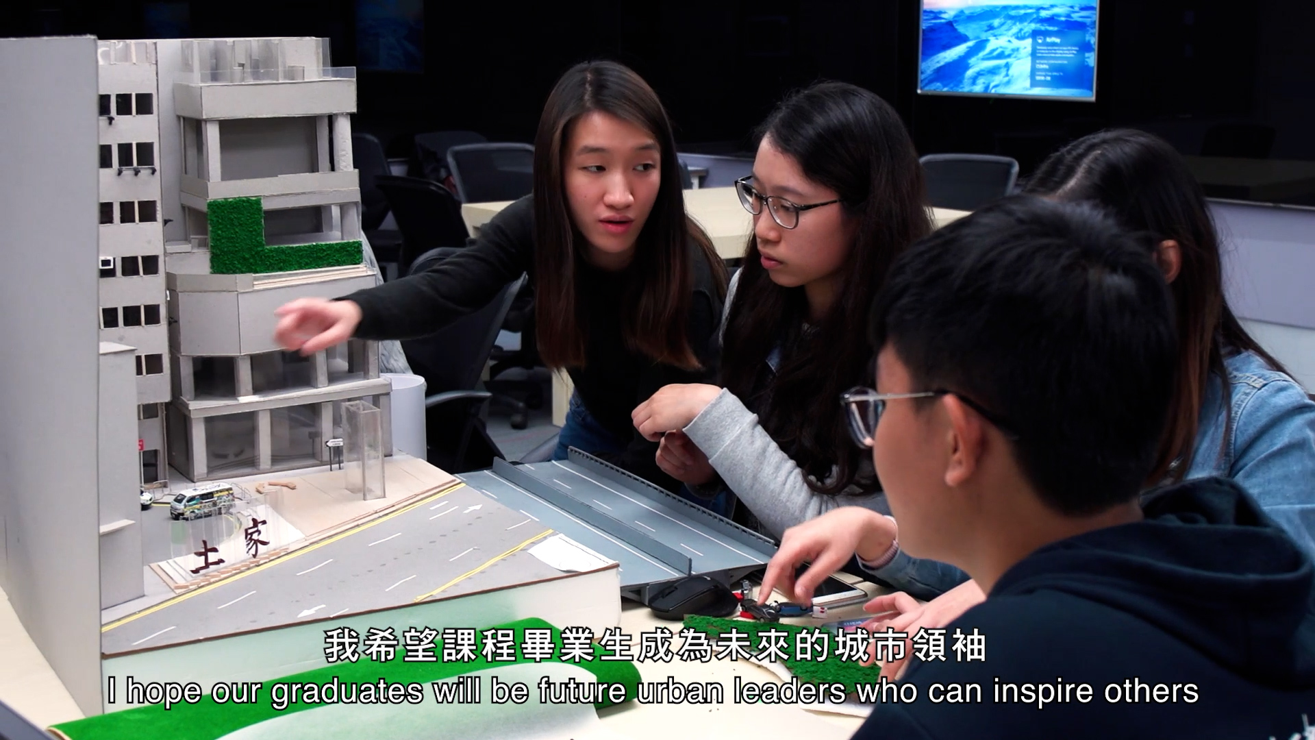 Four students sit around a table examining a scale model of a building and street in a classroom. One student points at the model while others observe. English and Chinese subtitles are visible at the bottom of the image.
