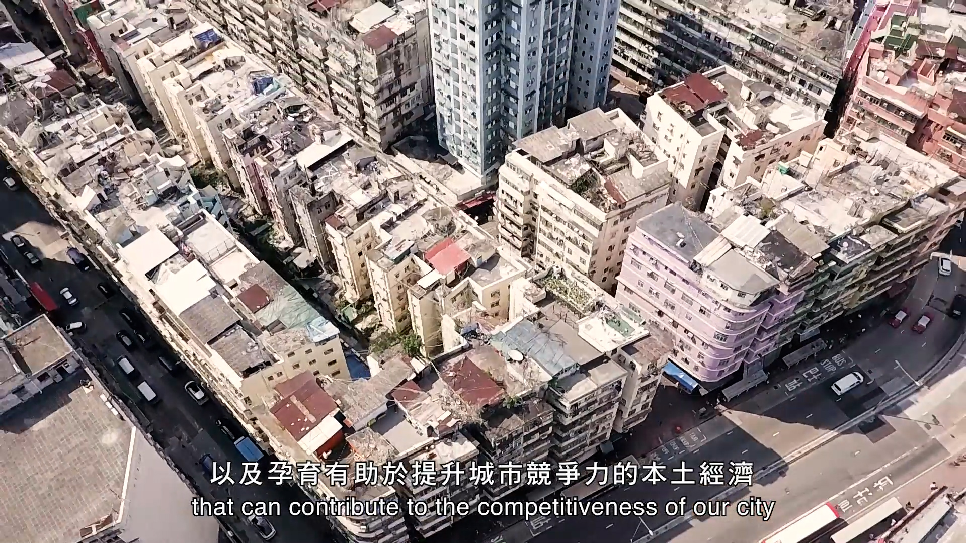 An aerial view of densely packed, weathered apartment buildings in an urban area, with subtitles in Chinese and English at the bottom of the image. The English subtitle reads: “that can contribute to the competitiveness of our city”.