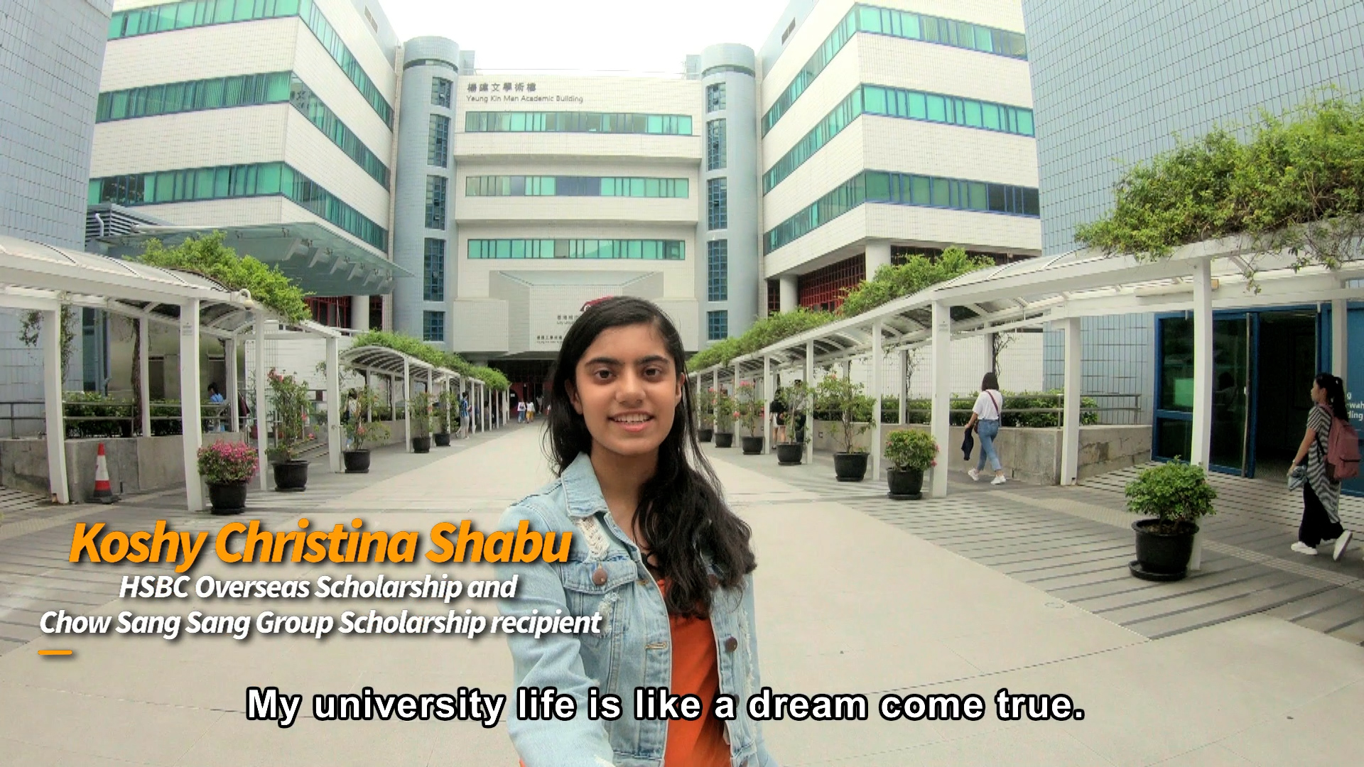 A young woman stands smiling outside a modern university building, taking a selfie. Text on the image introduces her as Koshy Christina Shabu, a scholarship recipient. Caption reads: "My university life is like a dream come true.