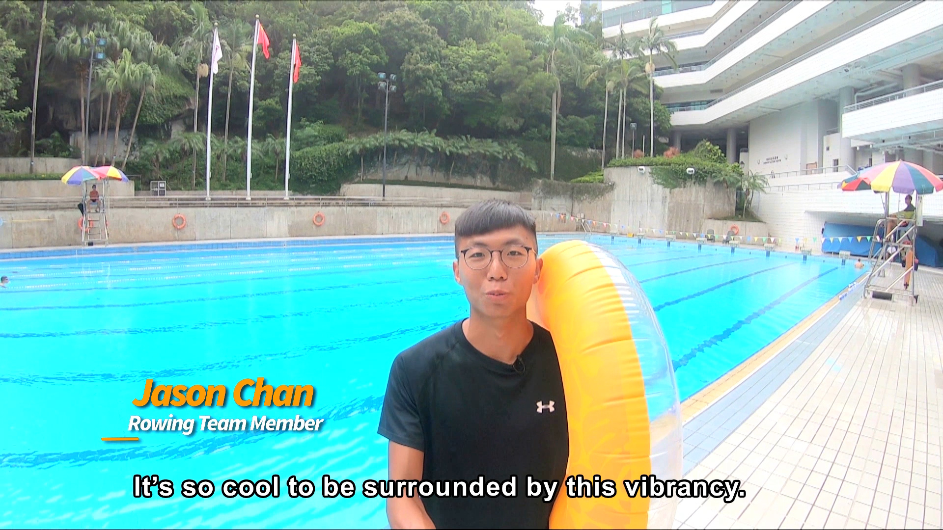 A young man in glasses stands by an outdoor swimming pool, holding a yellow inflatable ring. Onscreen text reads, “Jason Chan, Rowing Team Member. It’s so cool to be surrounded by this vibrancy.”.