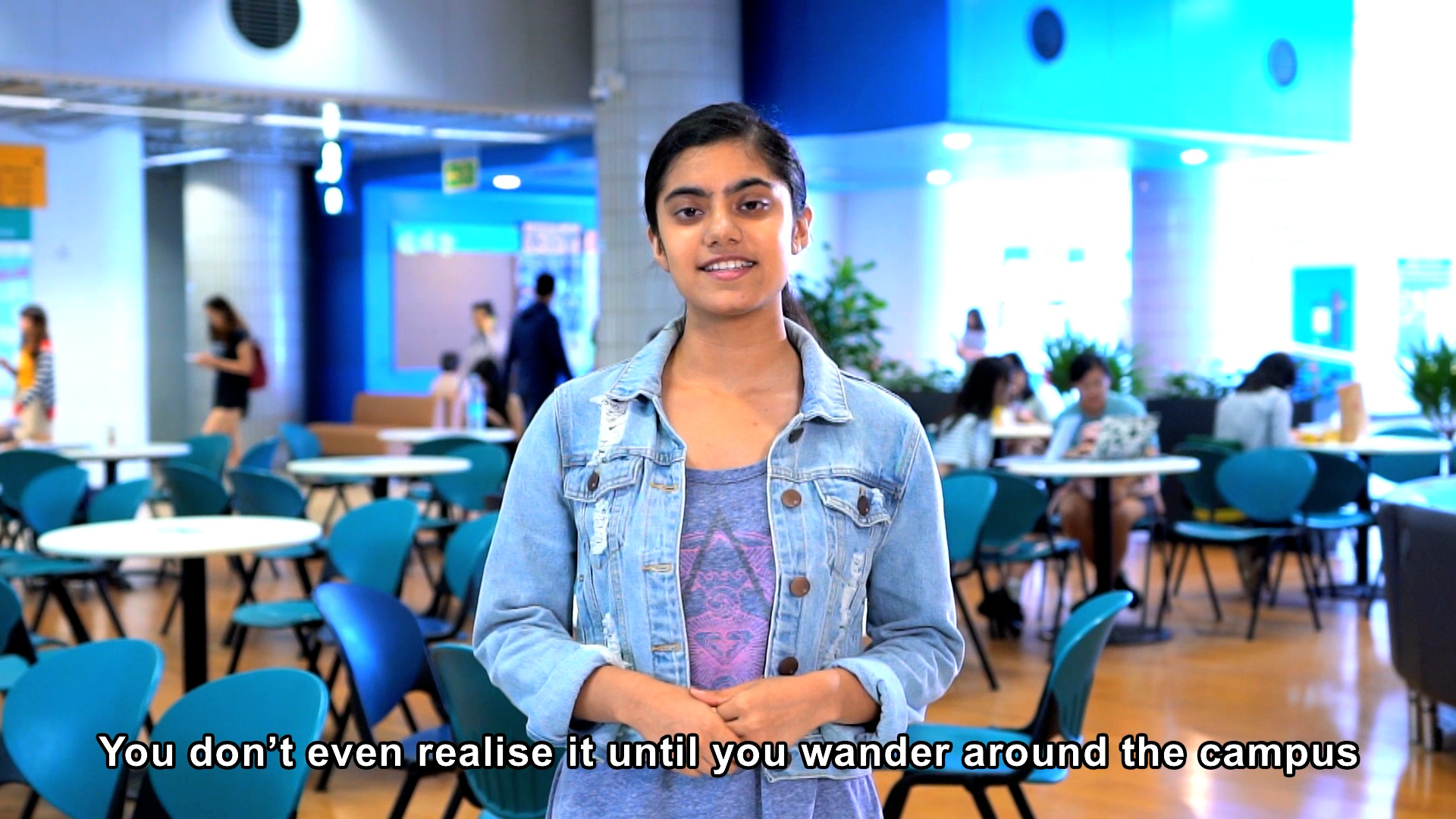 A young woman stands in a brightly lit campus cafeteria, speaking while wearing a denim jacket. Several people sit at tables in the background. Subtitle reads: "You don't even realise it until you wander around the campus.
